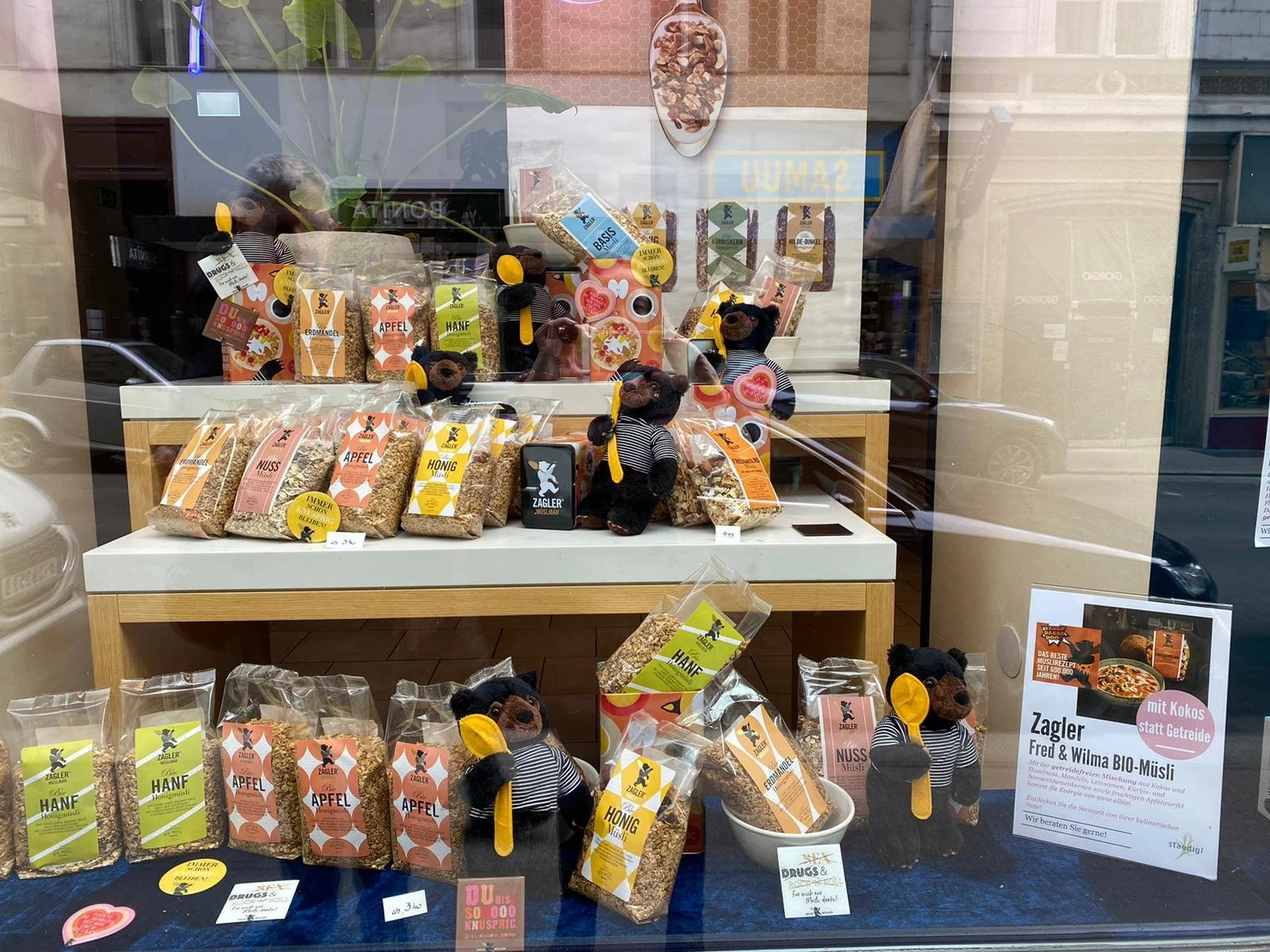 Schaufenster mit verschiedenen Bio-Müsli-Produkten, kleinen Teddyfiguren in Piratenkostümen, und einer Informationenstafel zum Bio-Müsli von Zagler, mit einem Carport und Gebäuden im Hintergrund.