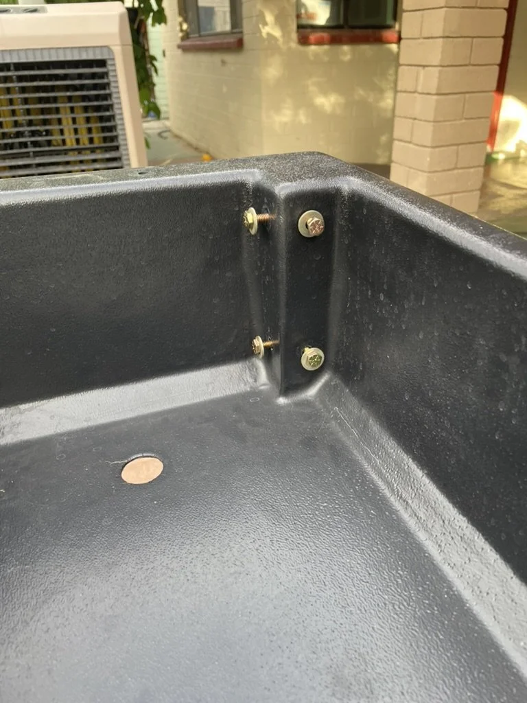 Close-up of a black metal surface with four screws, two of which are missing, and a penny on the surface. In the background, residential buildings and outdoor equipment are visible.