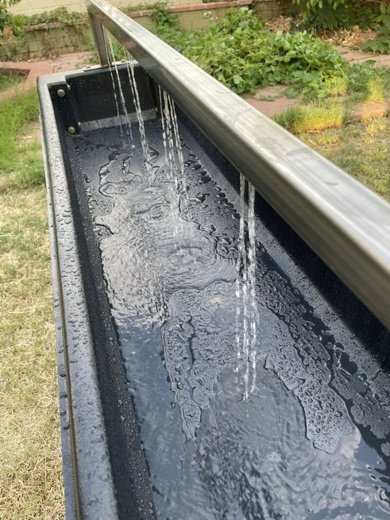 A black outdoor water trough with flowing water spilling from a metal pipe, set in a garden with green plants and grass.