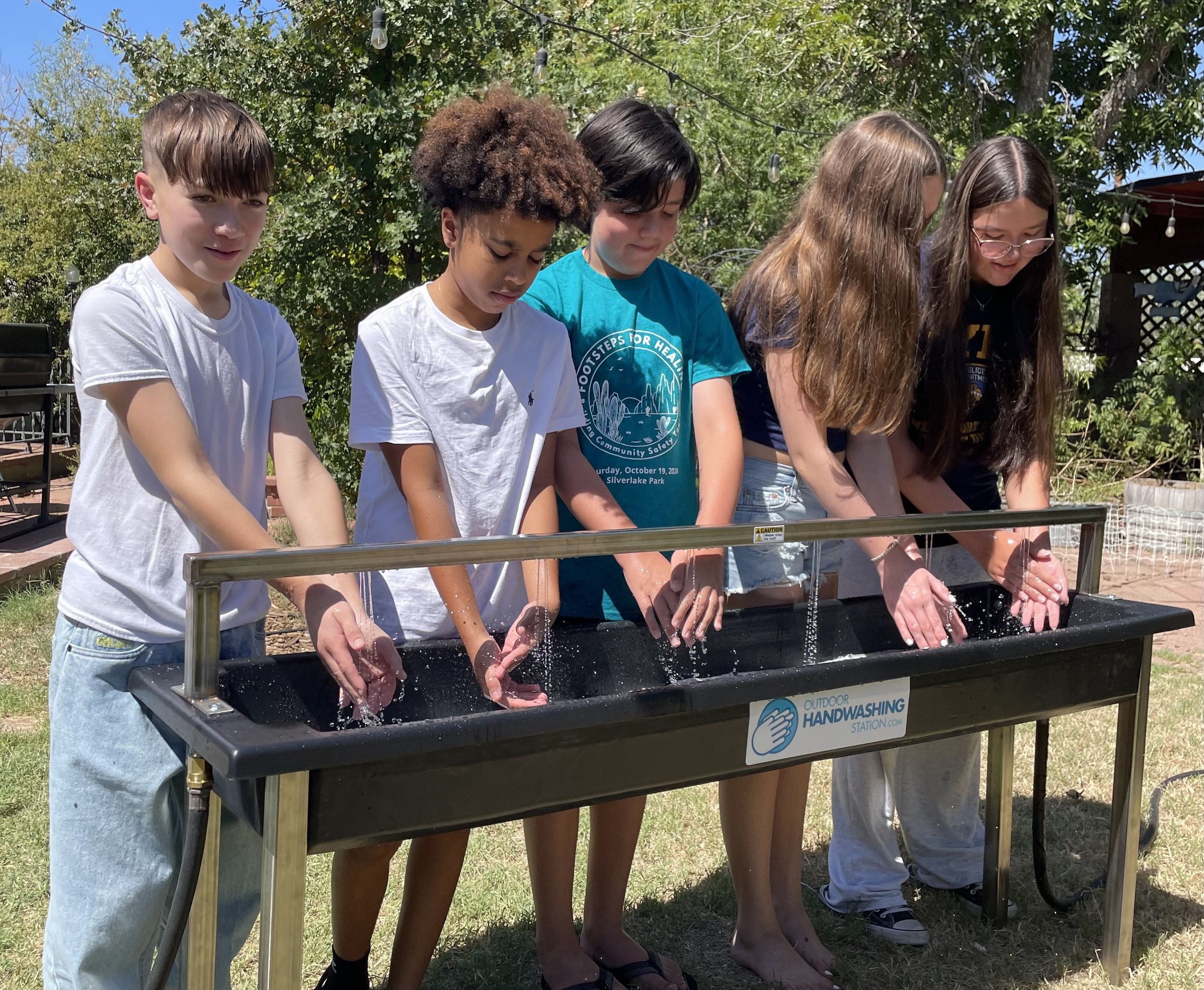 Middle and elementary school age children using a 6-person outdoor handwashing station on a lawn.
