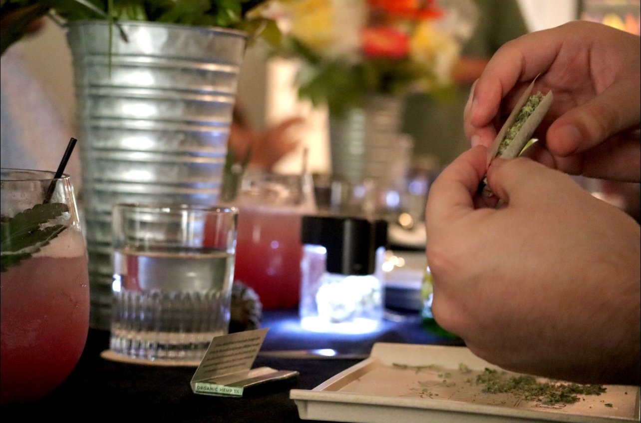 Person rolling a joint or cigarette with cannabis herb at a table with drinks and flowers.