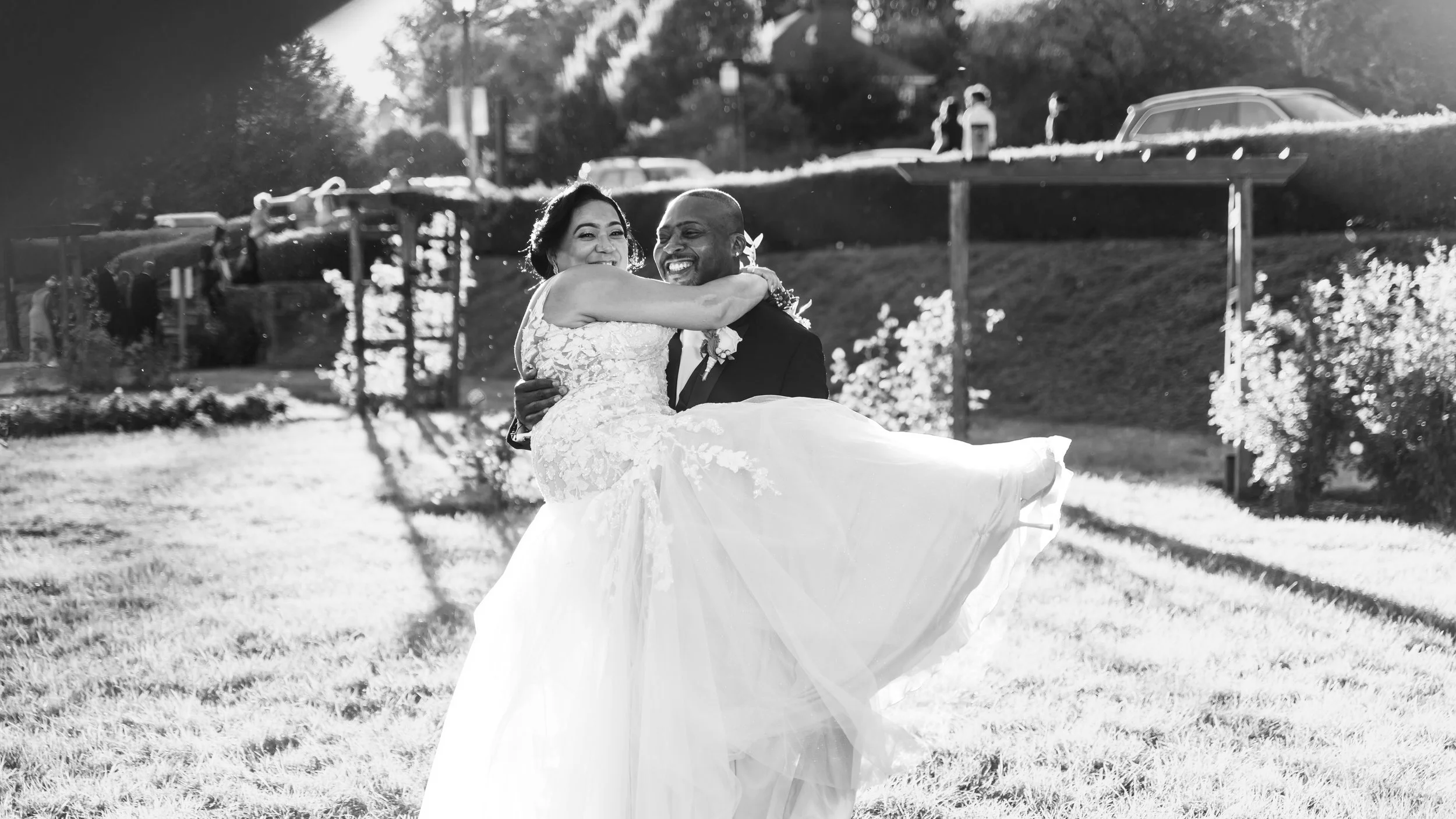 A bride and groom are dancing outside on a sunny day, with the groom lifting the bride in his arms. The scene is bright with sunlight and trees in the background.