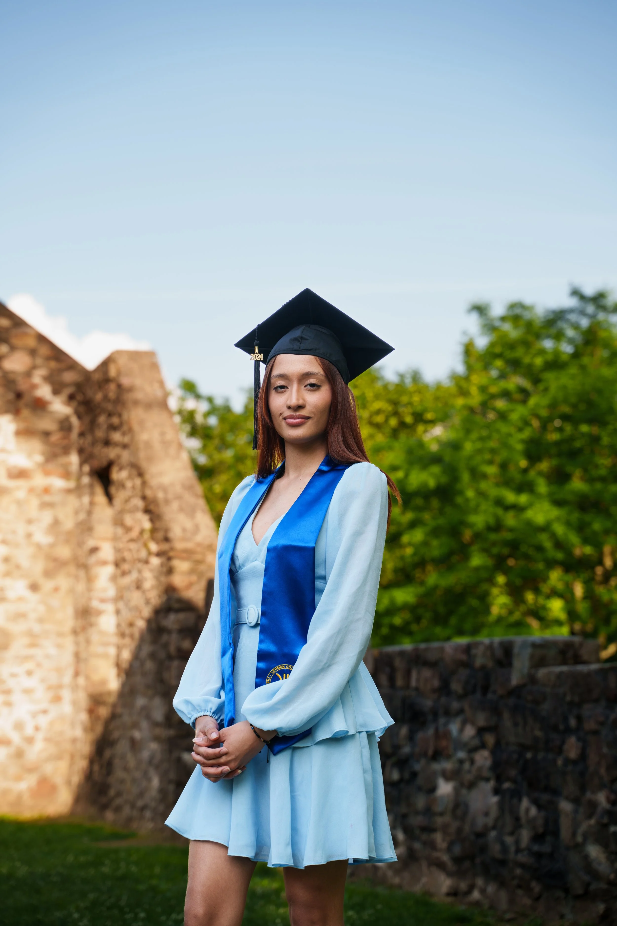 A young woman in a light blue graduation dress and a blue sash, wearing a black graduation cap with a gold tassel, standing outdoors near a stone structure and green trees, posing for a graduation photo.