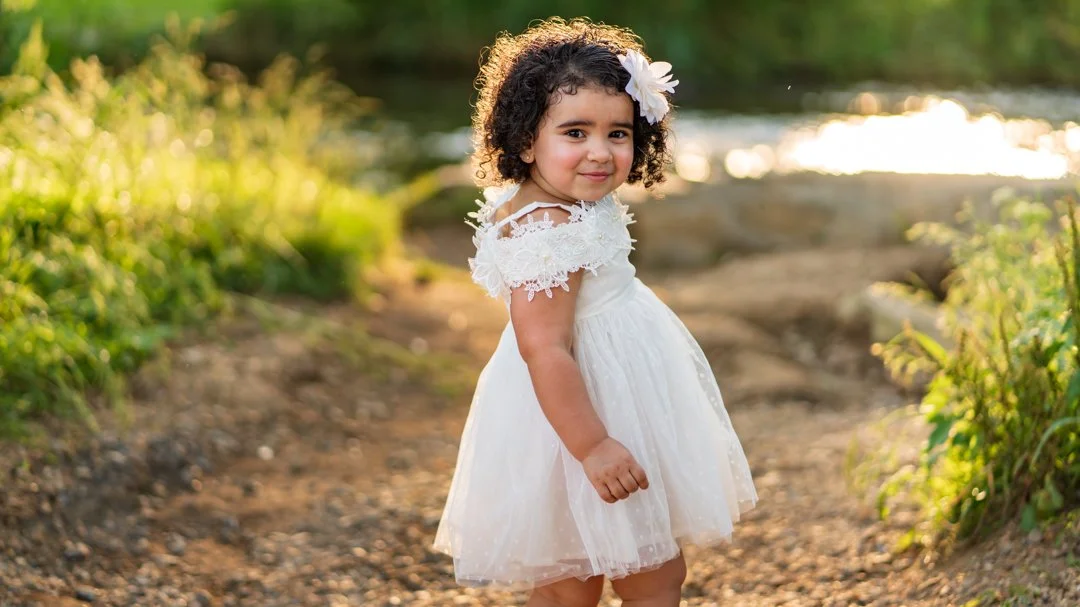 A young girl with curly hair and a flower in her hair, wearing a white dress, standing on a dirt path near a body of water with greenery, during sunset.