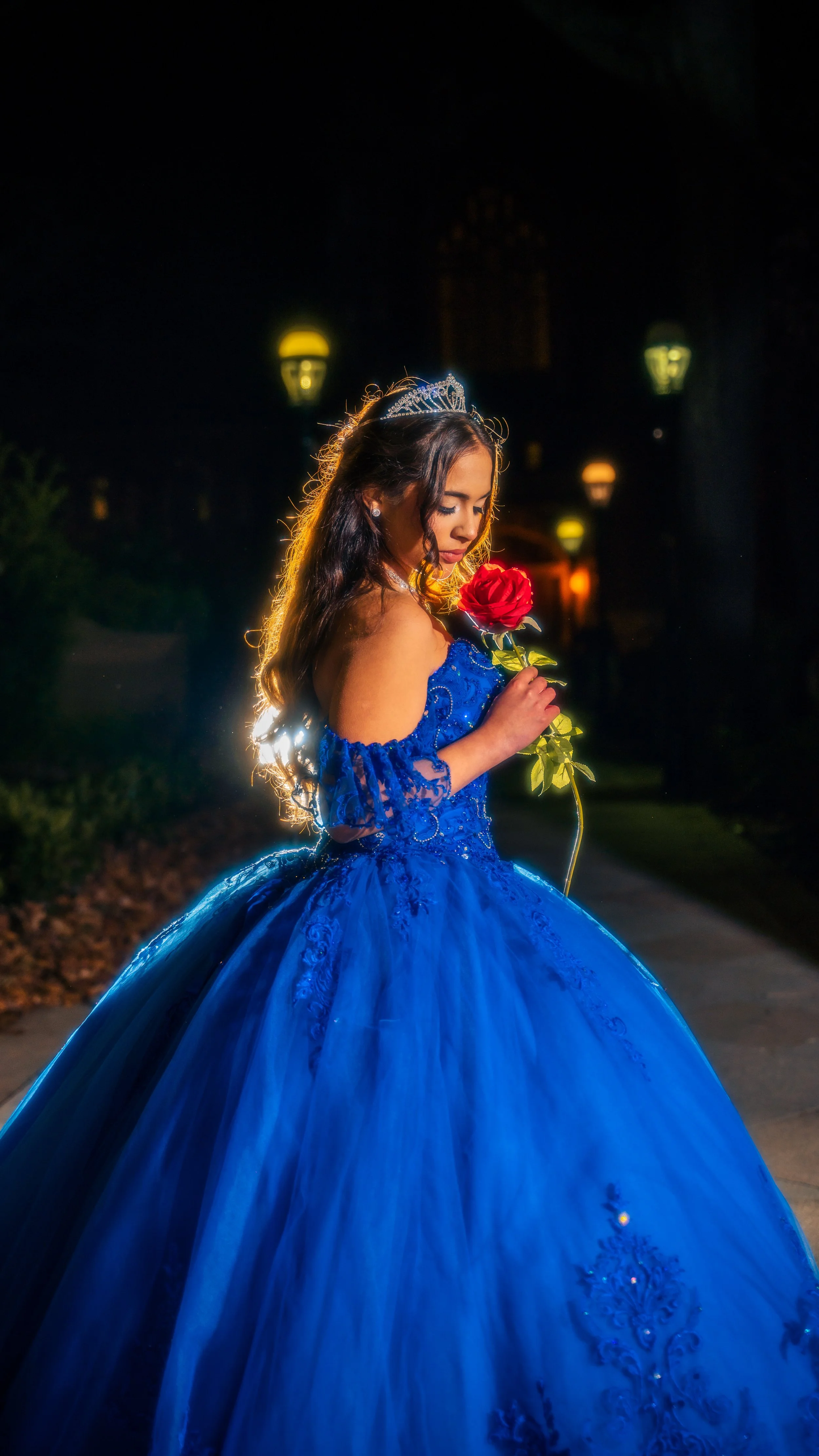 A woman in a blue gown and tiara holding a red rose at night, with street lamps glowing in the background.