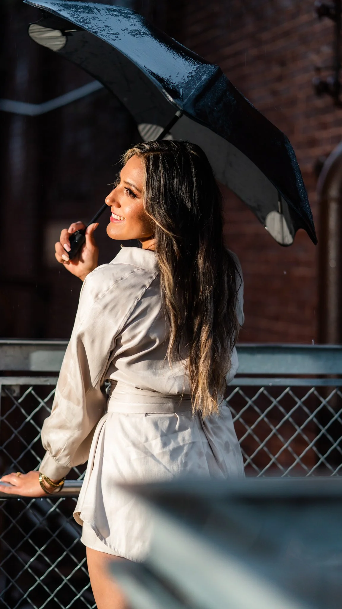 A woman with long wavy hair is holding a black umbrella and smiling while looking to the side.