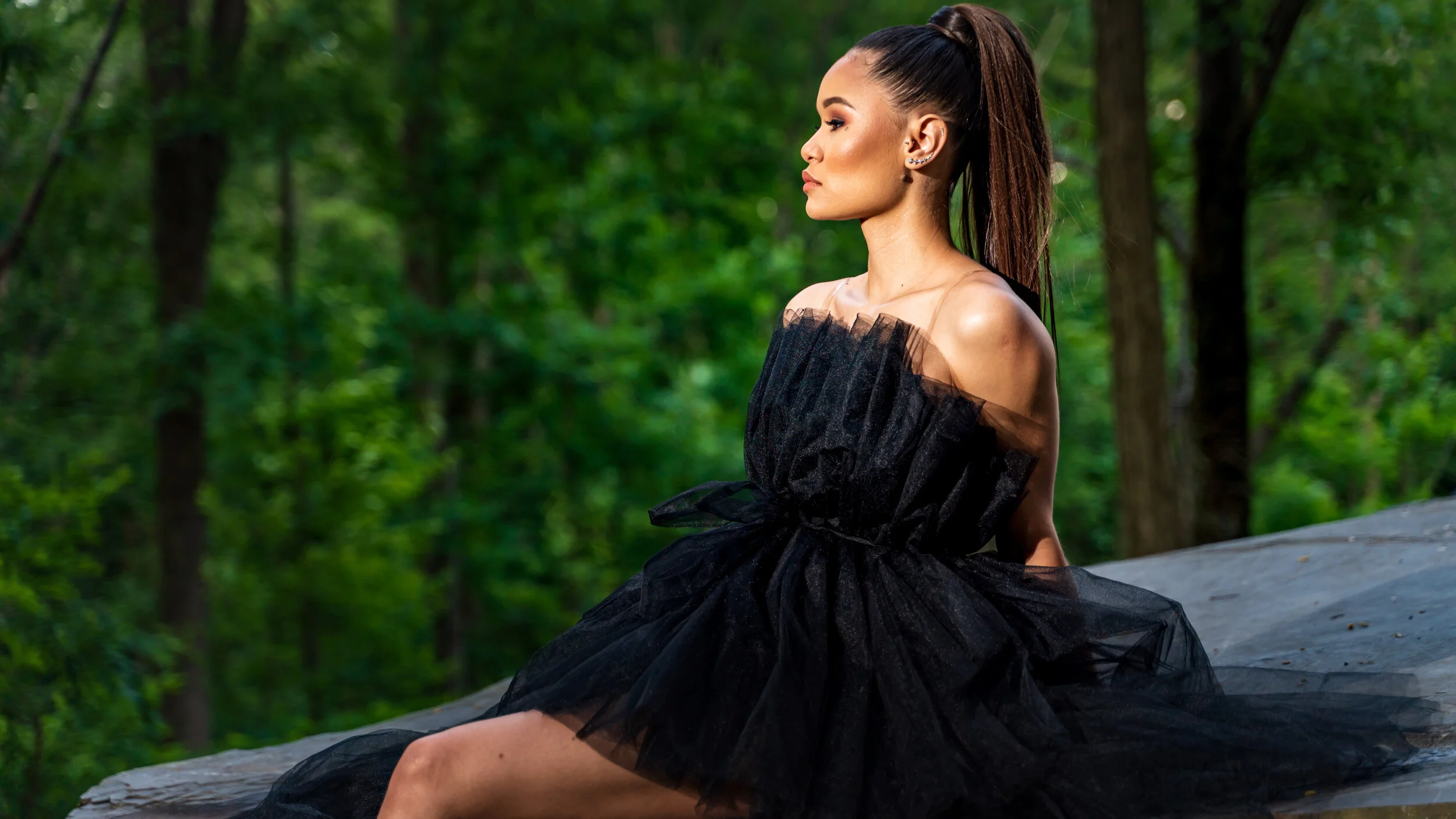 A woman in a black off-shoulder dress sitting outdoors on a flat rock, with a background of green trees.