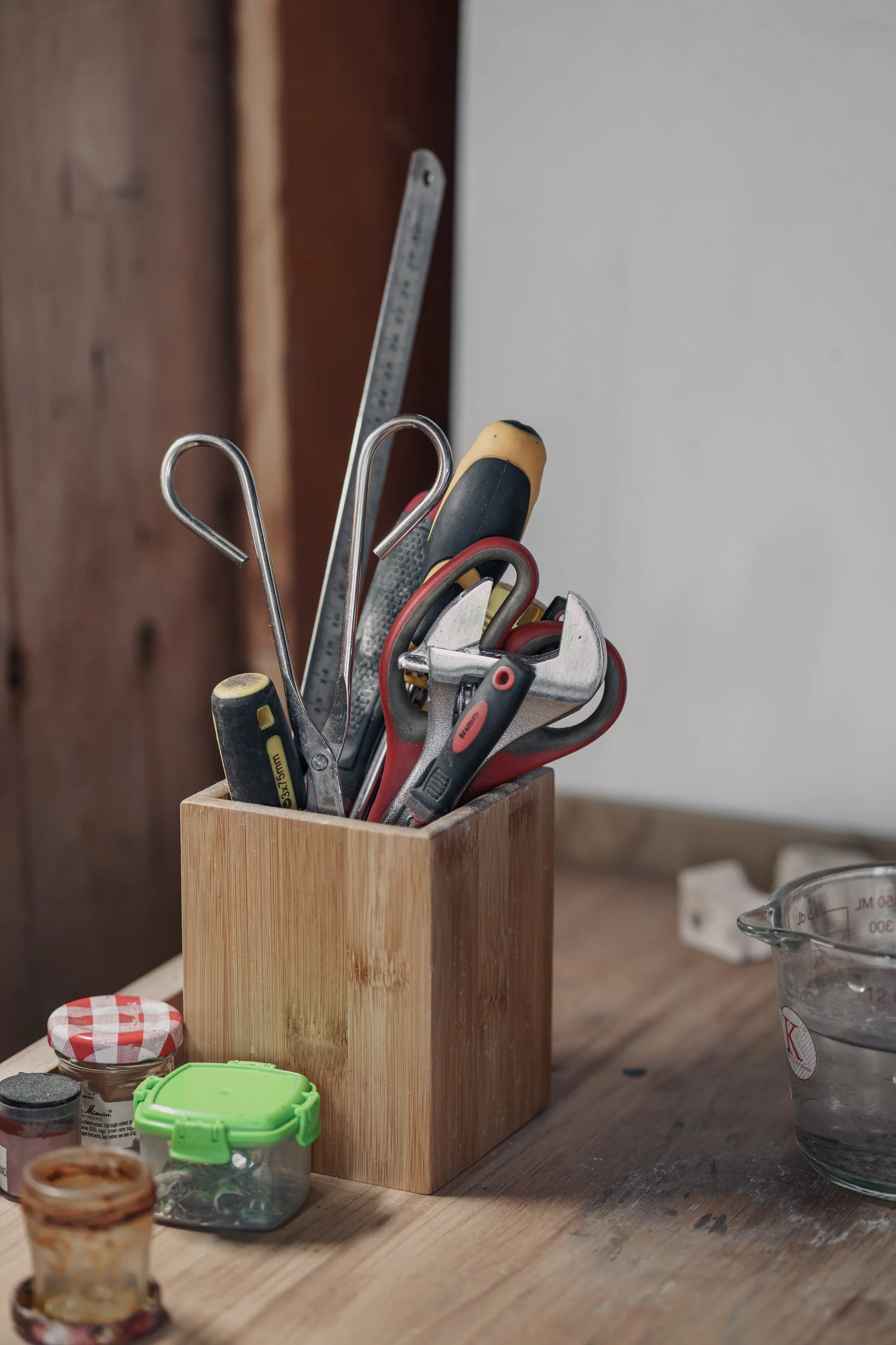 A wooden block holding various tools, including pliers, scissors, and a screwdriver, on a workbench with small containers and a measuring cup nearby.