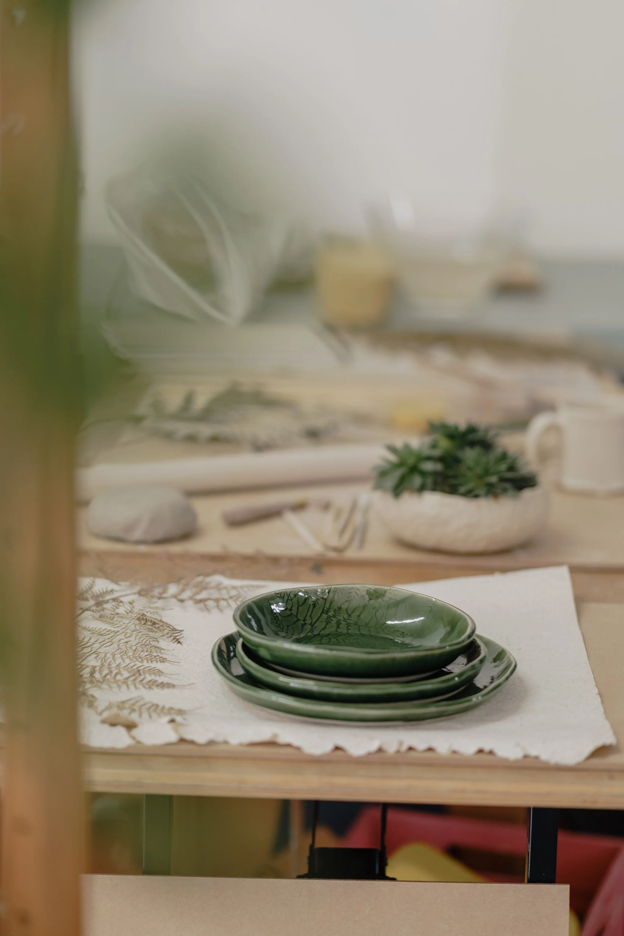 Green ceramic plates on a table with a white cloth napkin and fern design; blurred background with decorative items and small potted plant.