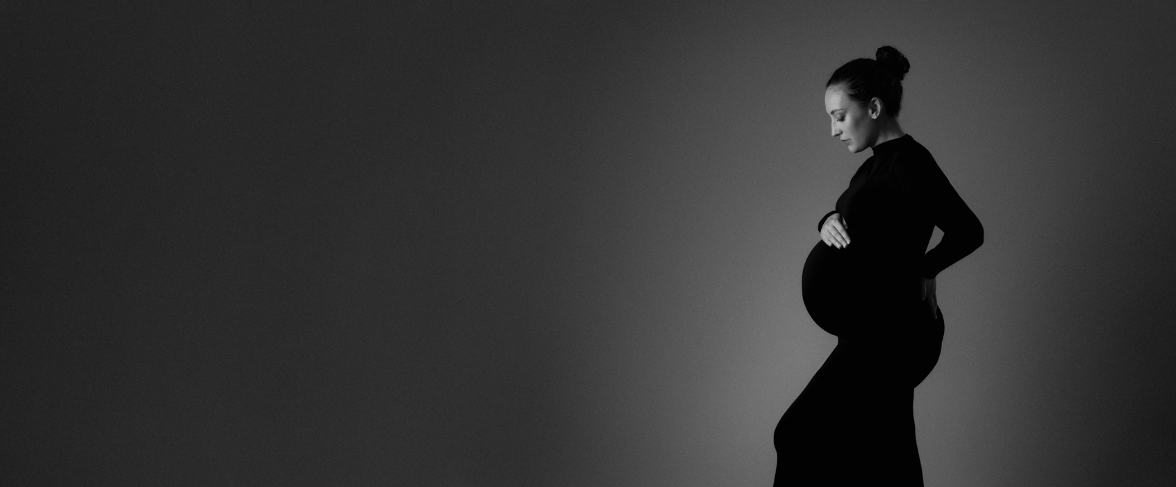 A pregnant woman standing elegantly holding belly in black long dress. The photo is in black and white.