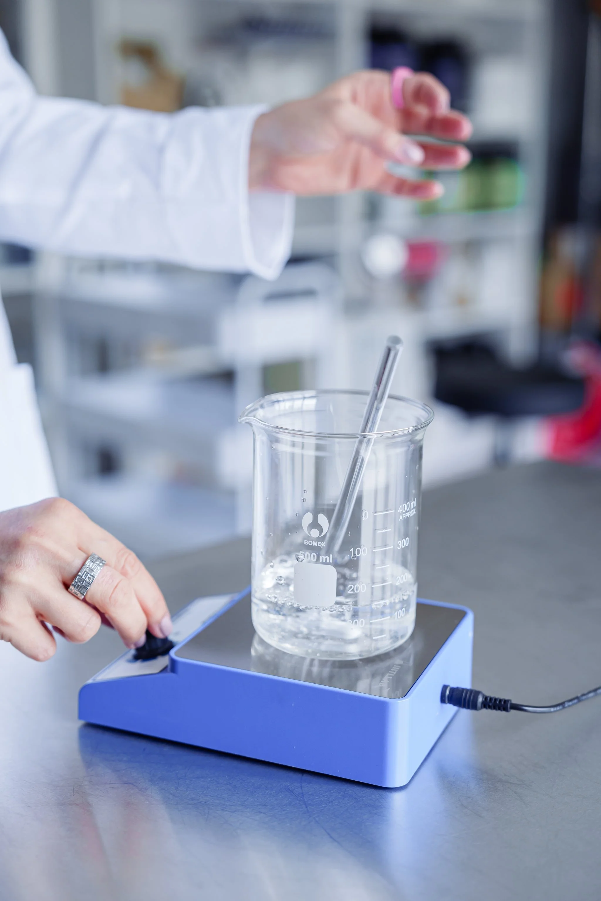 A laboratory setting with a close-up of a person adjusting a magnetic stirrer while a glass beaker containing liquid and a stir bar is placed on the device.