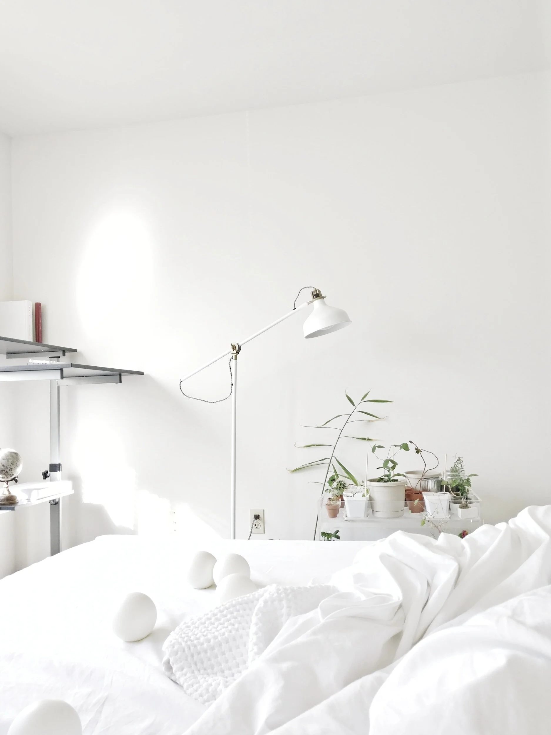 Minimalist bedroom with white bedding, scattered white decor items, a white floor lamp, and potted plants on a shelf against a white wall.
