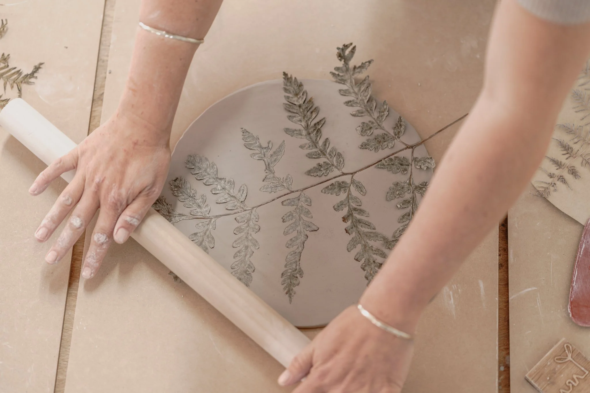 Person using a rolling pin to press fern leaves into clay