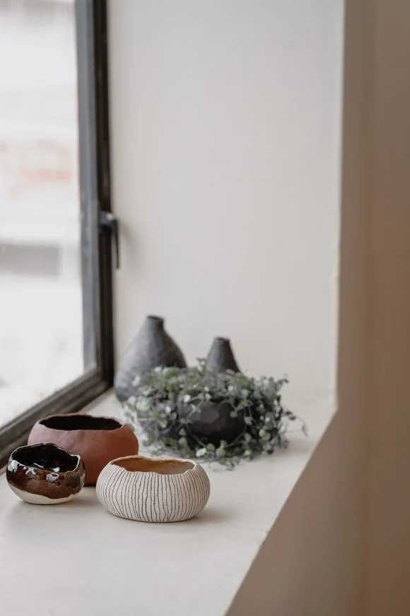 A windowsill with decorative ceramic bowls and black vases, with green foliage around the vases.