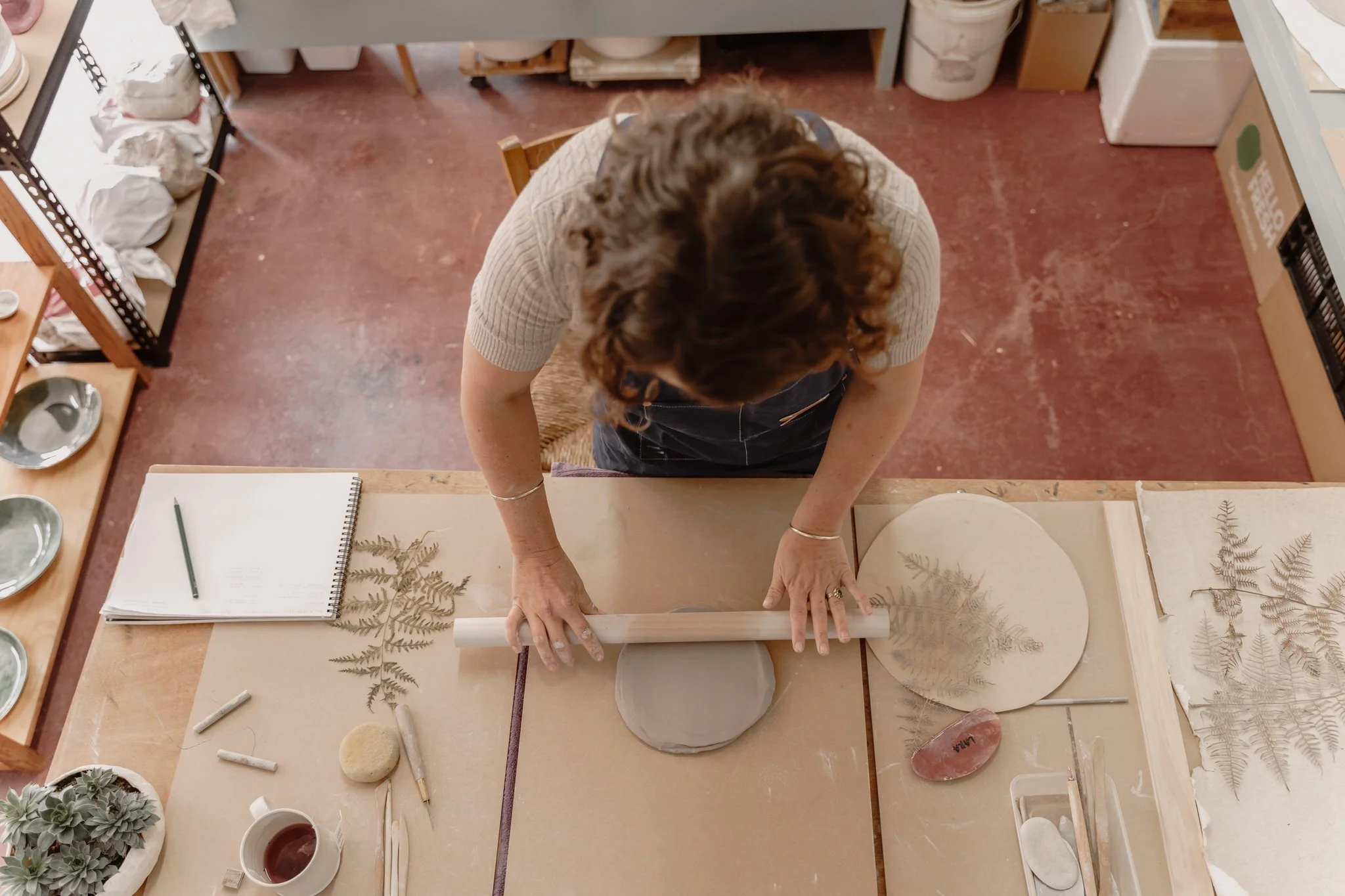 Overhead view of a person rolling clay on a wooden table in a pottery studio, surrounded by tools, a notebook, and fern leaves for imprinting.