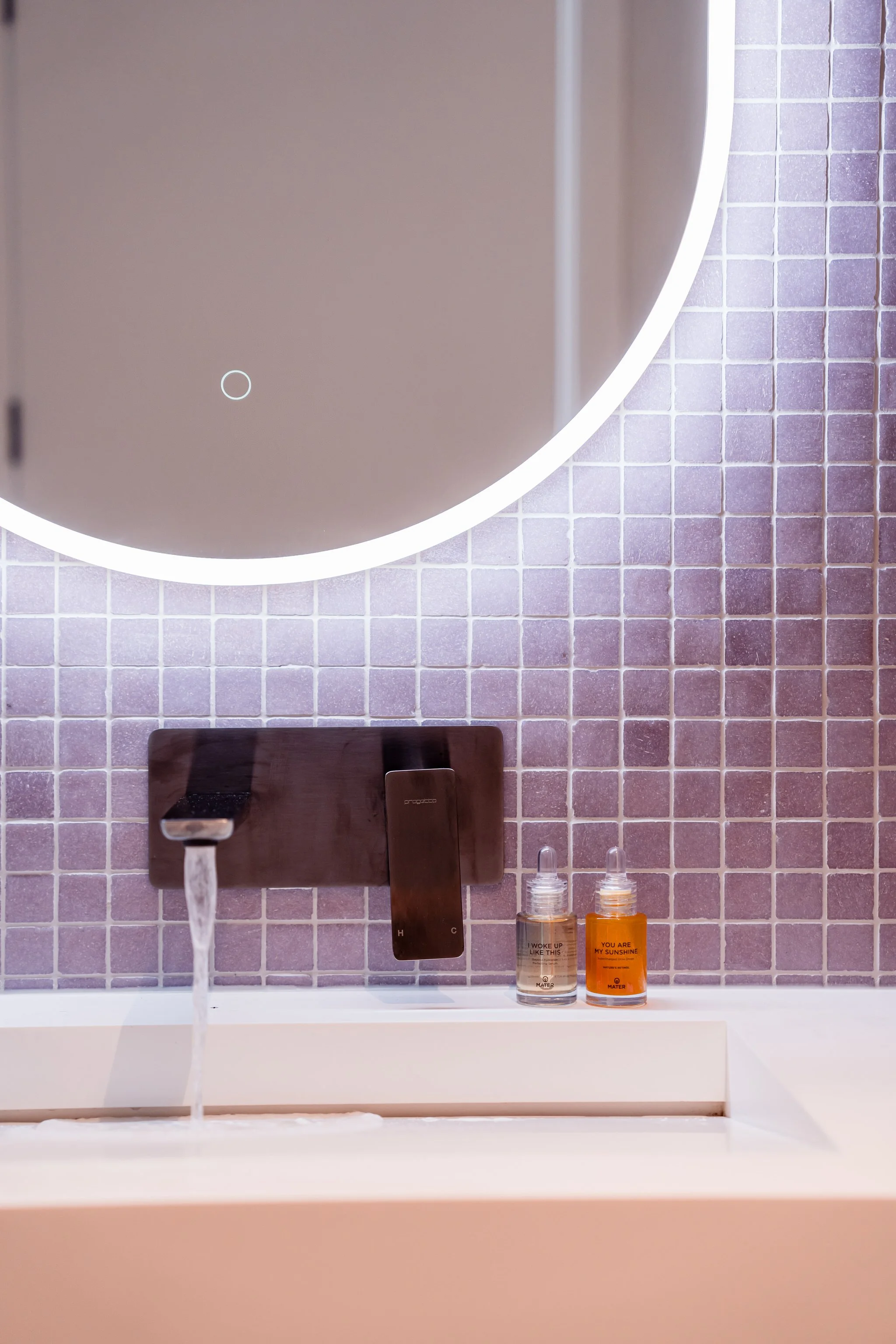 Modern bathroom sink with running water faucet, round illuminated mirror, purple mosaic tile backsplash, and small bottles of toiletries.