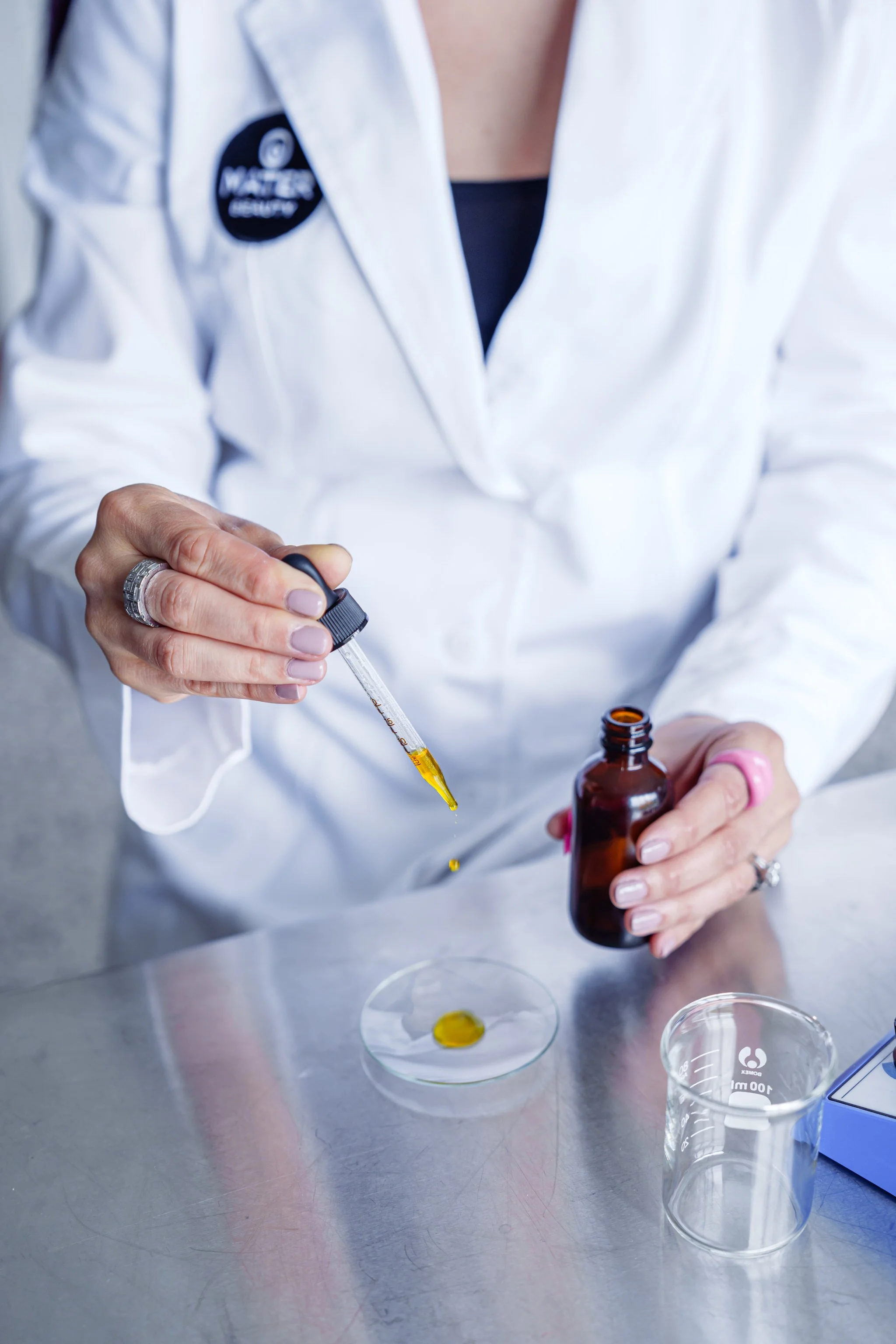 Person in lab coat using a dropper to transfer liquid from a brown bottle to a petri dish on a laboratory table.