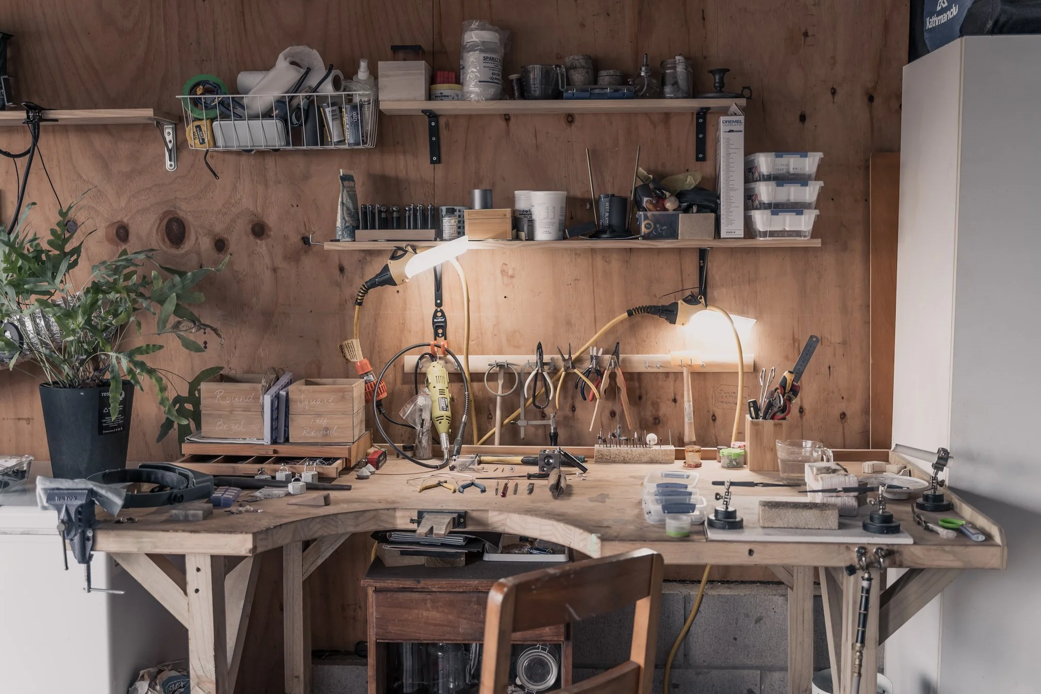 A cluttered workbench in a workshop with tools, materials, and a plant.