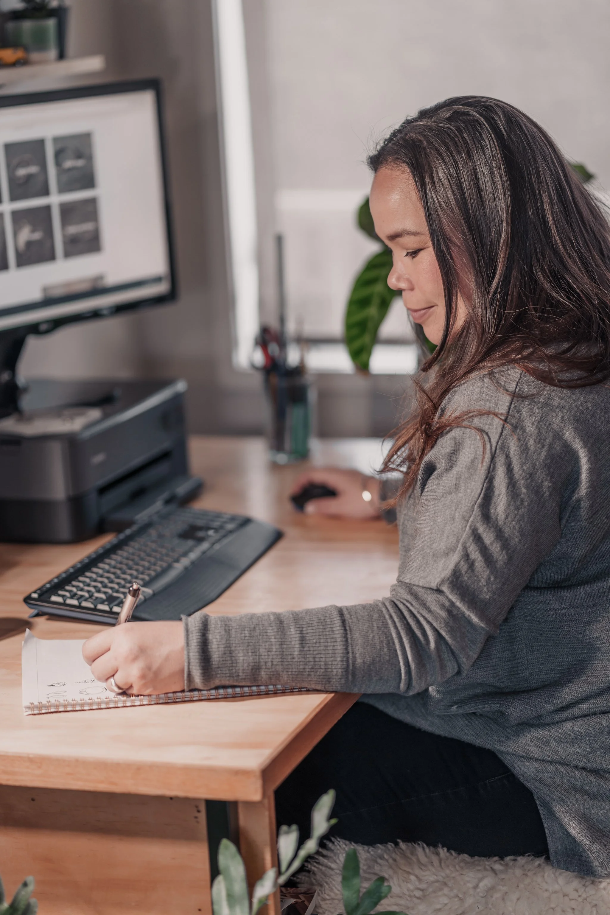 Woman working at a desk, writing in a notebook, with a computer and keyboard in front of her.