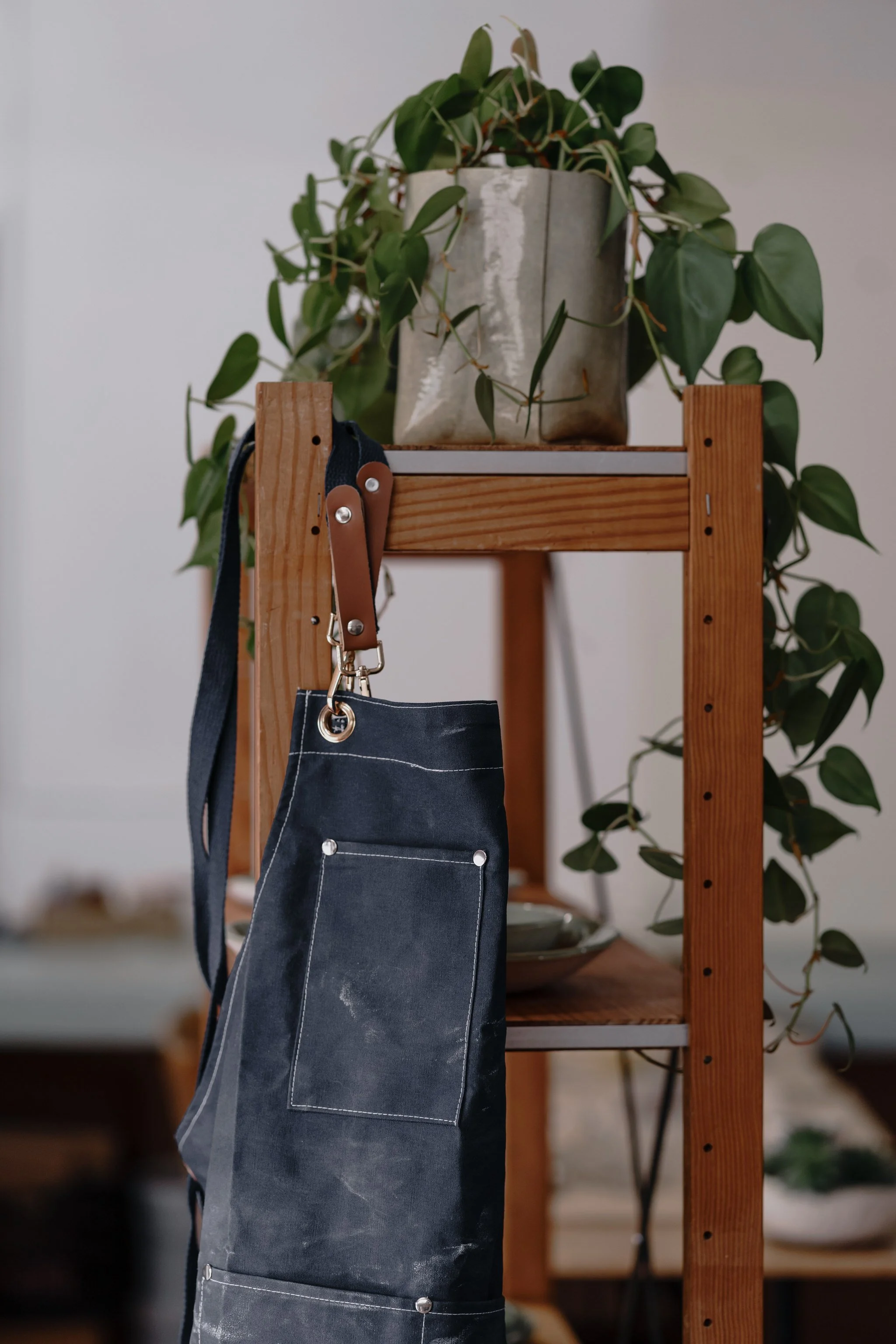 Canvas apron with leather straps hanging on a wooden shelf next to a potted plant with green leaves.