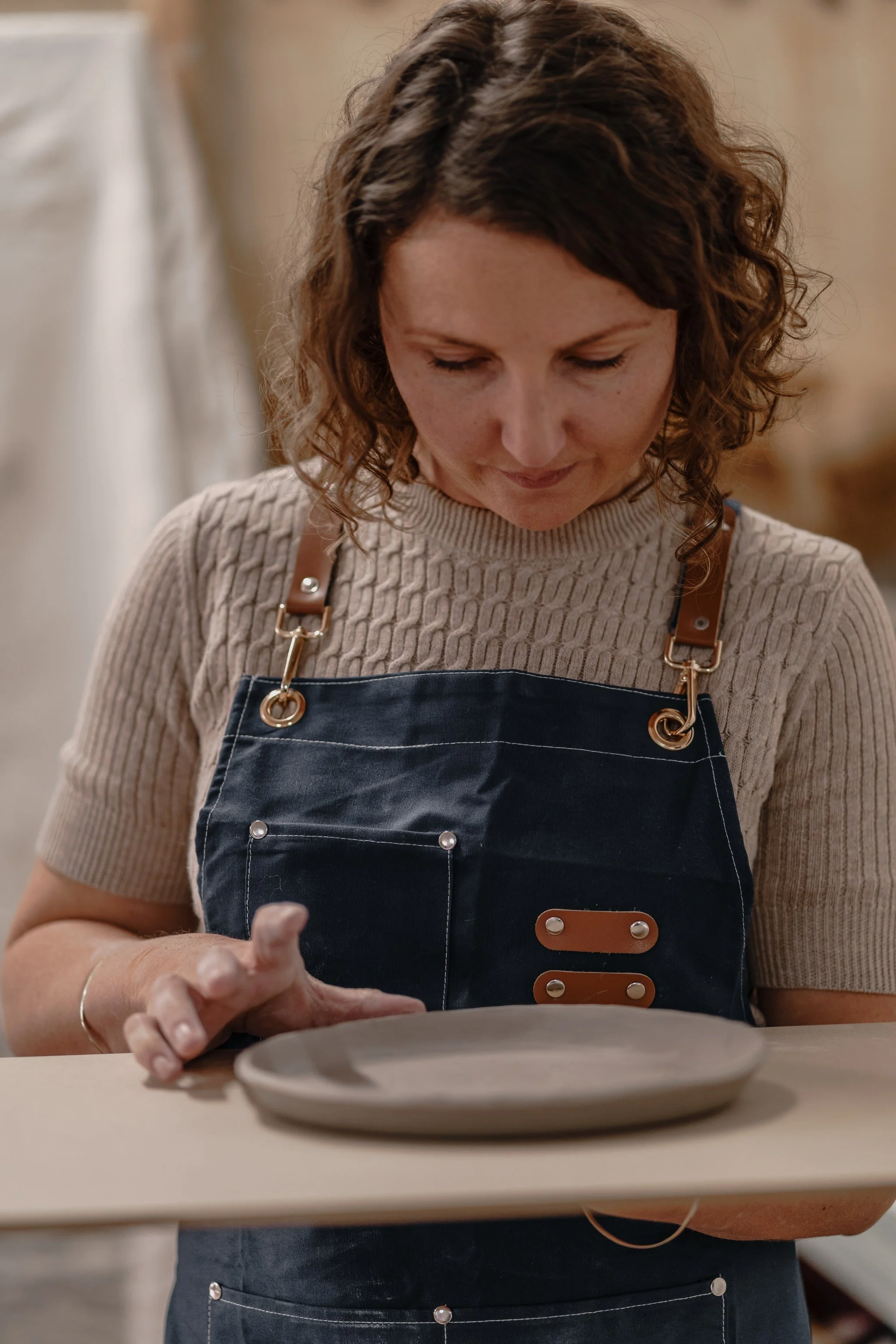 Woman shaping a clay plate in a workshop, wearing a blue apron and beige sweater.
