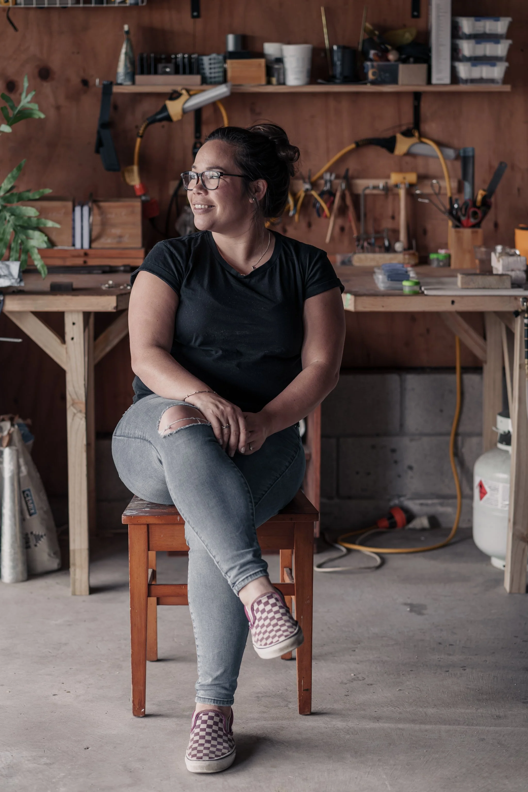 Person sitting on a stool in a workshop, surrounded by tools and equipment, wearing a black t-shirt, jeans, and checkered shoes.