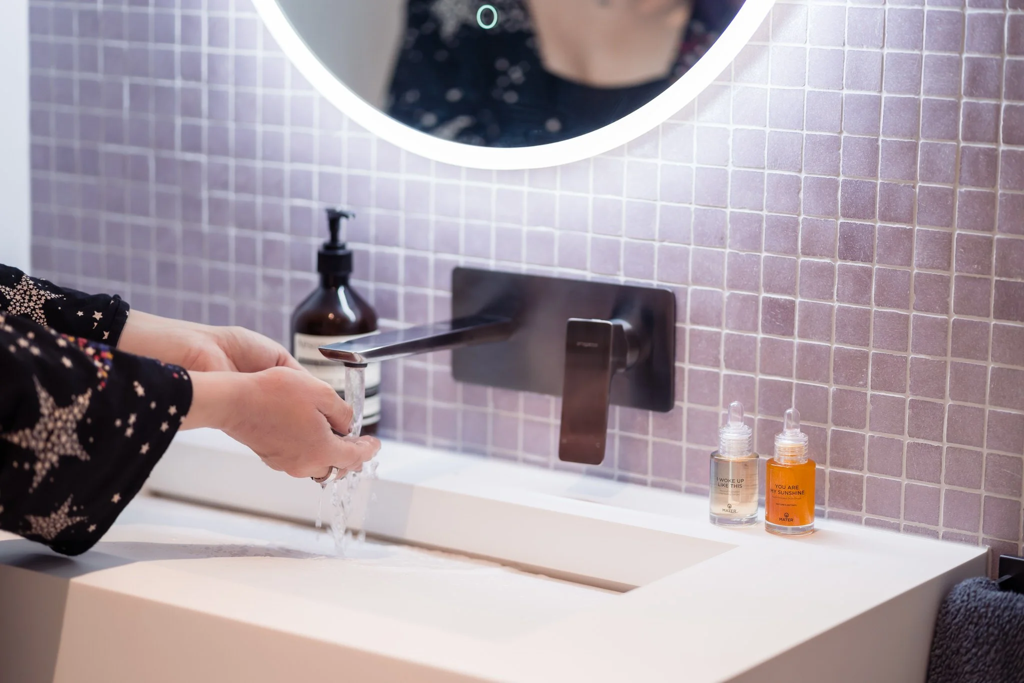 Person washing hands under a modern faucet with a backlit mirror and bottles on a tiled bathroom counter.