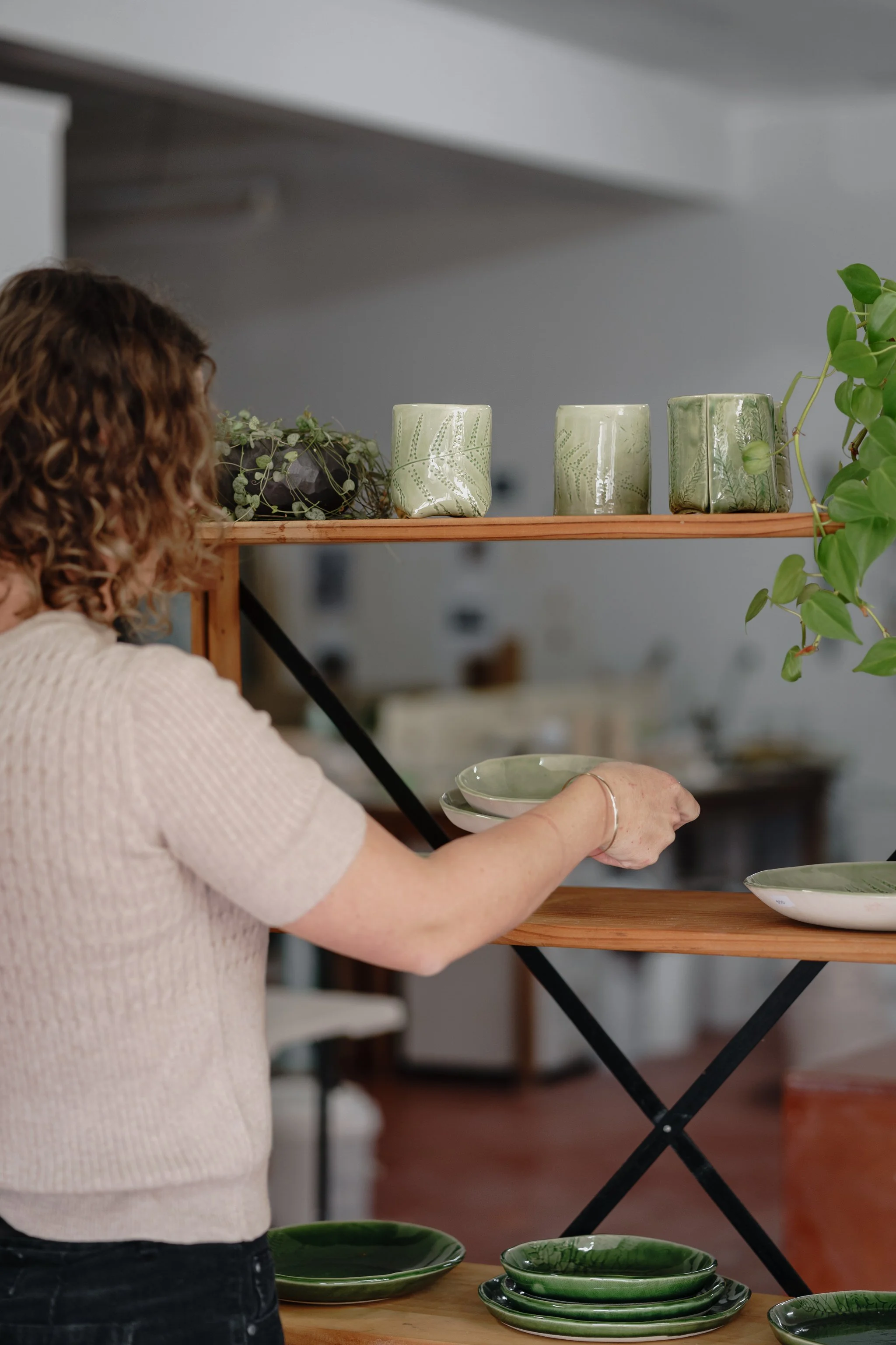 Person arranging green ceramic dishes and mugs on a wooden shelf with plants.