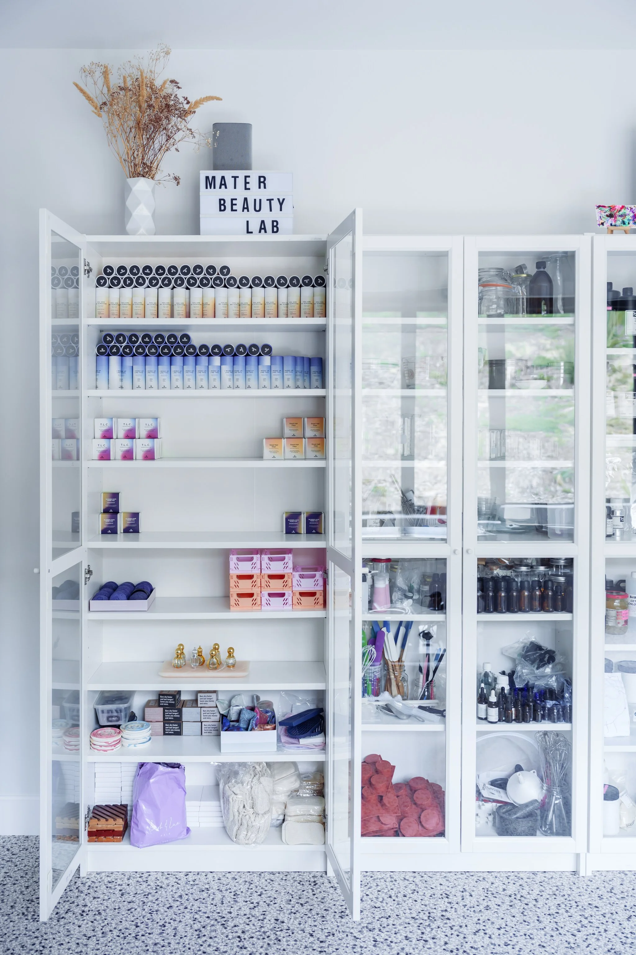 A white cabinet filled with neatly arranged cosmetic products, including bottles, boxes, and containers of various colors and sizes. The top shelf features a sign labeled 'Mater Beauty Lab' and a vase with dried flowers. The cabinet is in a room with