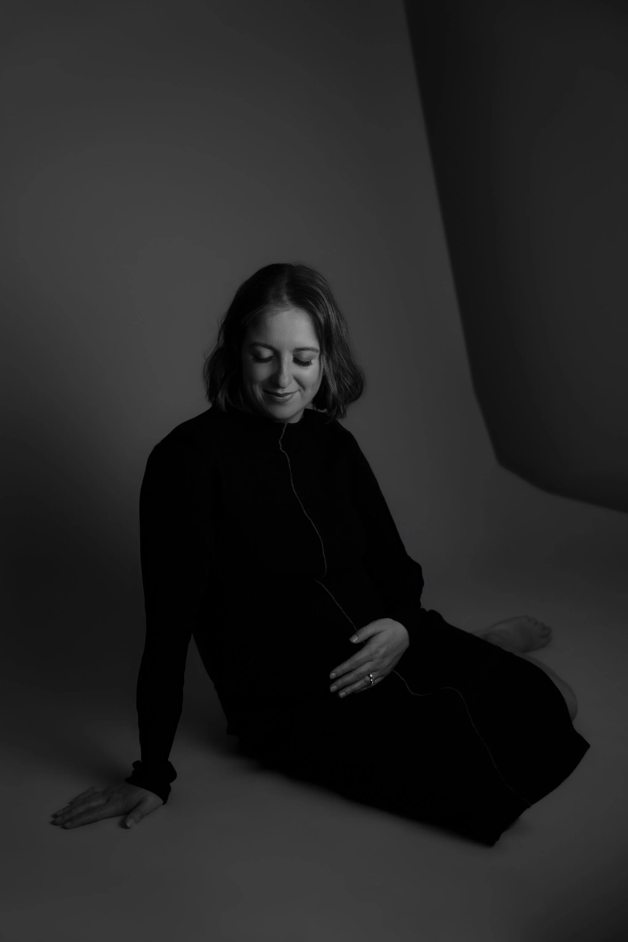 Black and white photograph of a pregnant woman in a seated pose, looking down and smiling gently, with her hands resting on her belly.