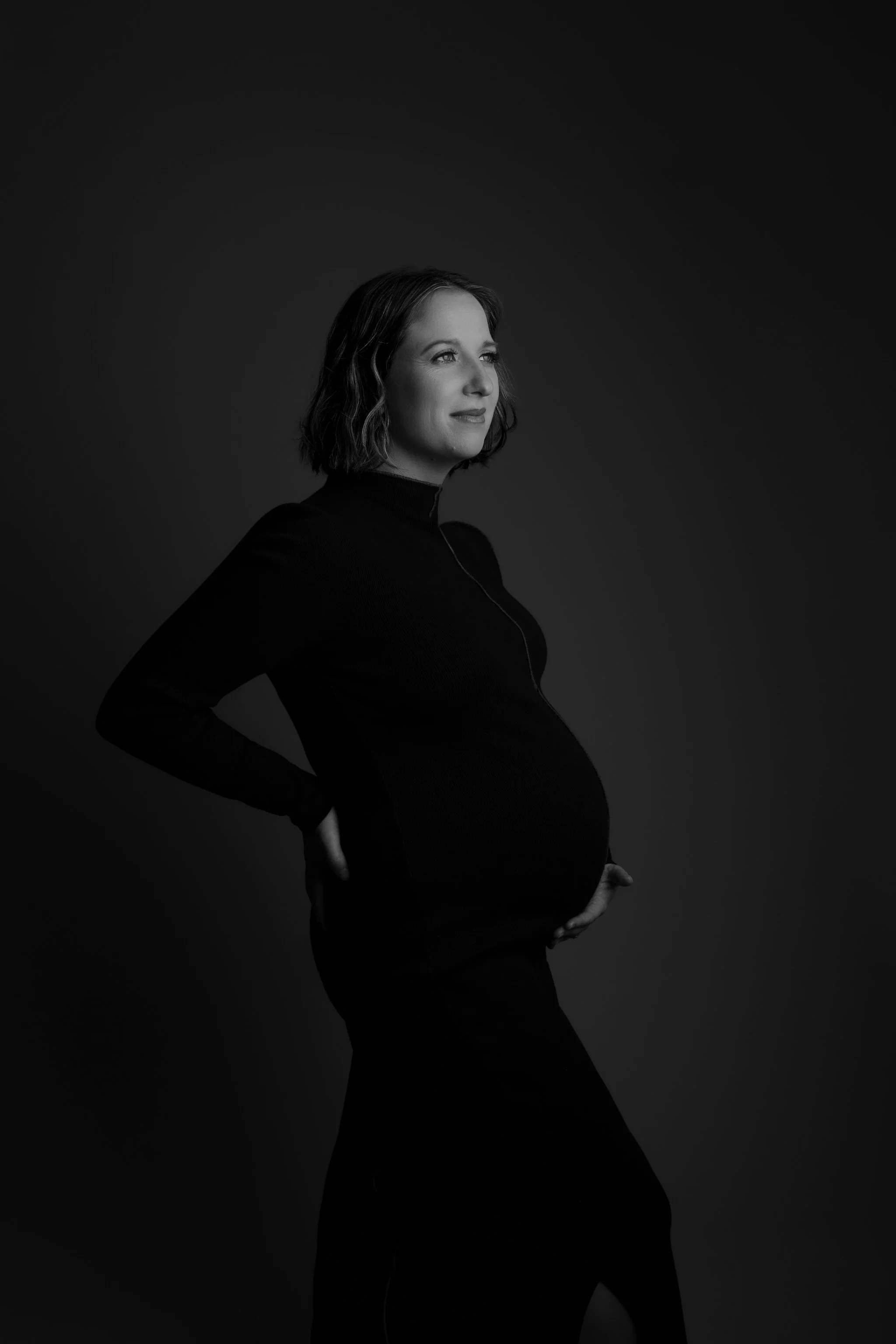 Black and white photo of a pregnant woman wearing a long sleeve top, standing with one hand on her hip and the other holding her belly, smiling softly and looking to the side.