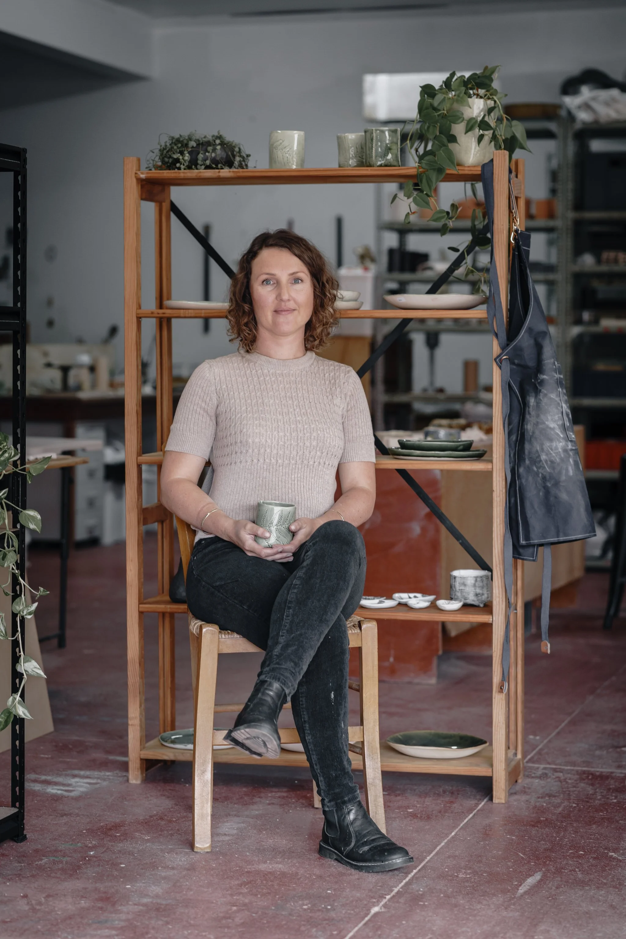 Woman sitting in a pottery studio holding a ceramic mug, with wooden shelves displaying various pottery items and plants.
