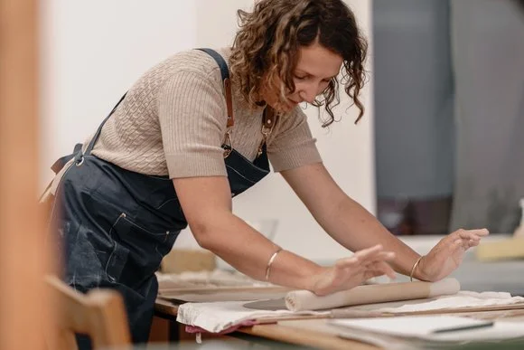 Woman using a rolling pin on a worktable, wearing a beige shirt and dark apron, in a craft or kitchen setting.