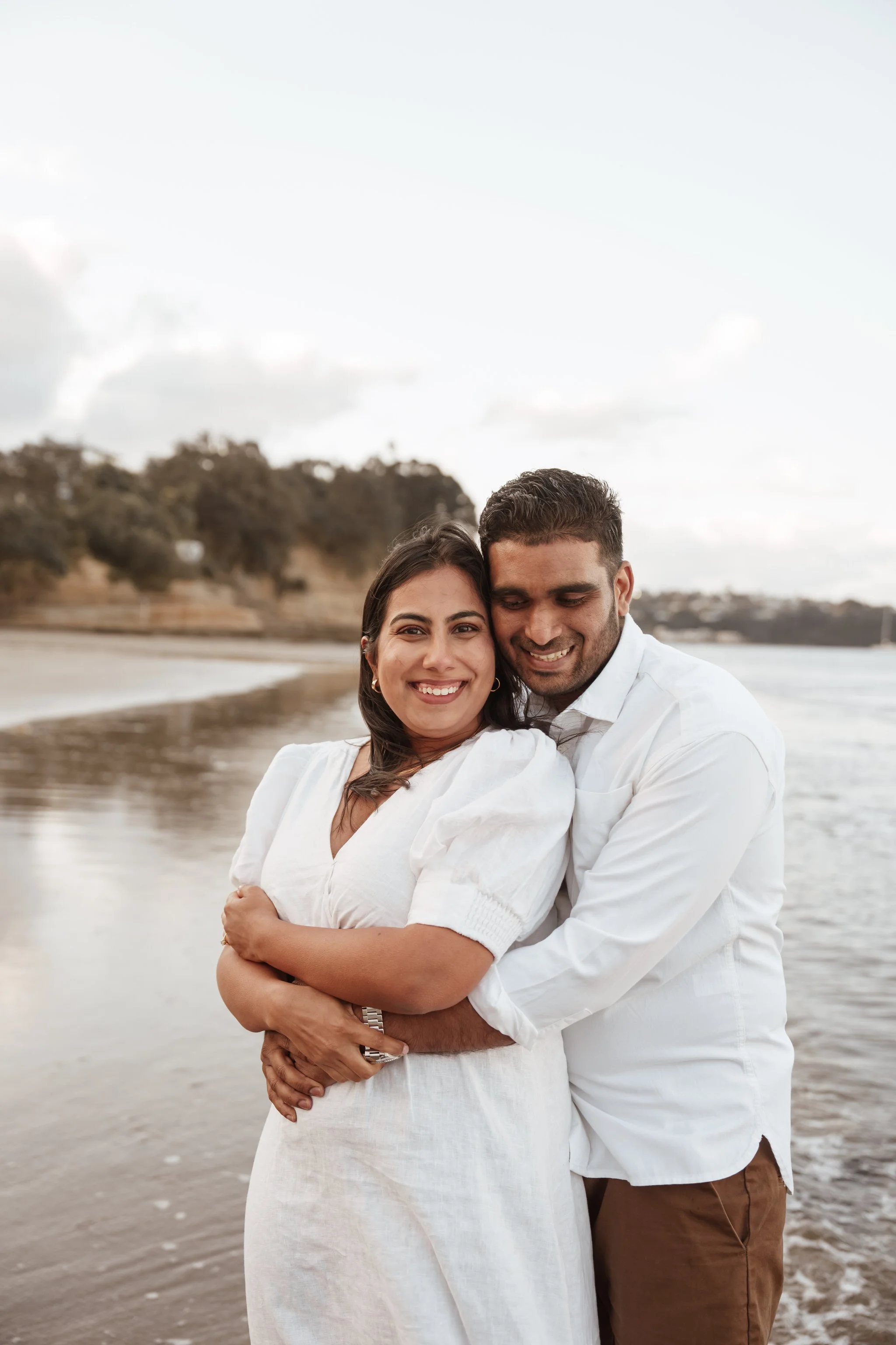 A woman in a white dress smiles while leaning on a man in a brown shirt, standing outdoors with trees in the background.