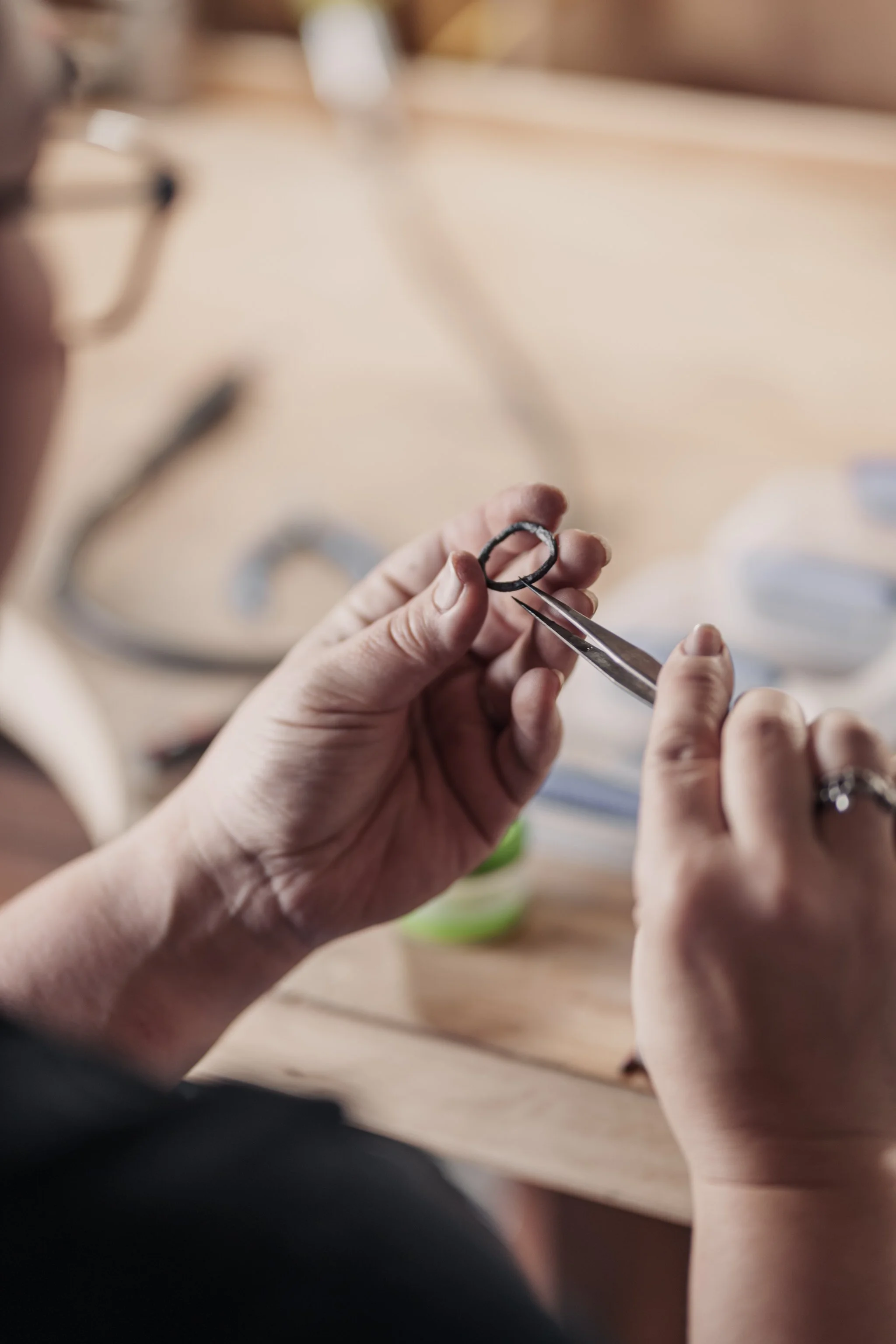 Person holding a small ring with tweezers at a workbench, close-up of hands.