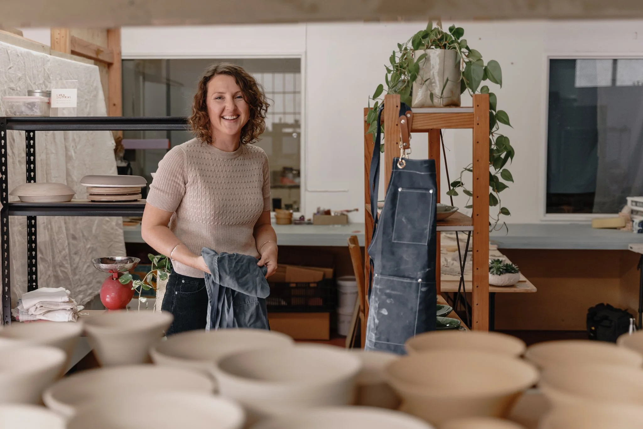 Woman in a pottery studio standing near shelves with pottery and a hanging apron, smiling, holding cloth, potted plant in background.