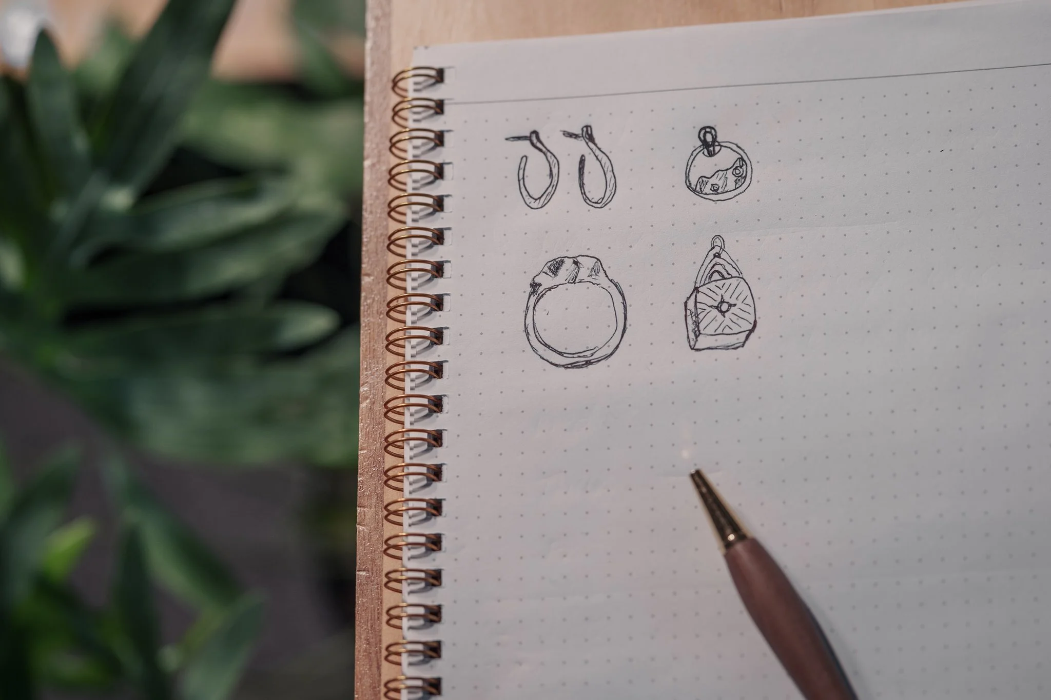 Sketches of jewelry designs including earrings, a pendant, and rings on a spiral-bound notebook with a pen on the side, set against a blurred green foliage background.