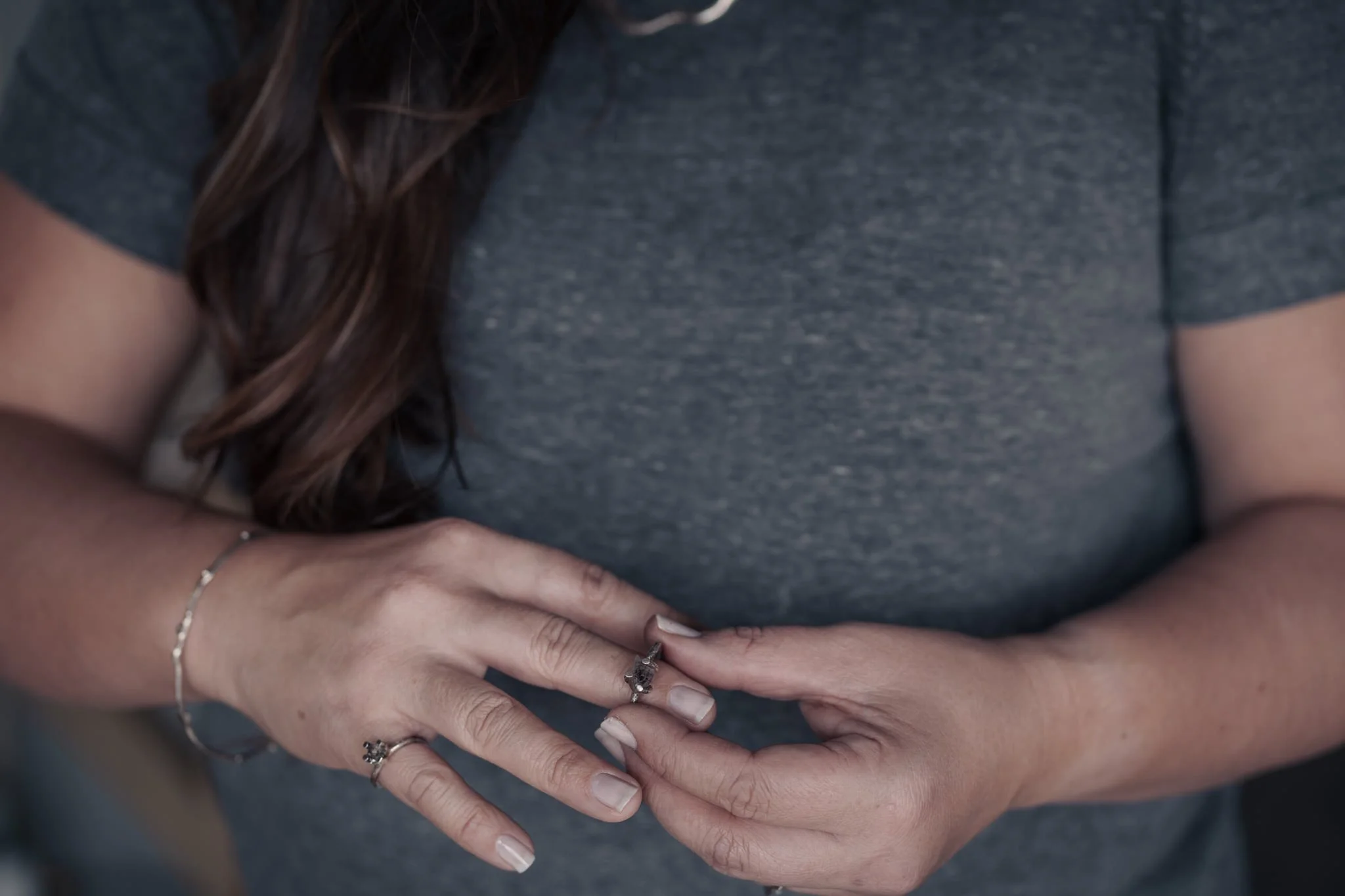 Person in gray shirt adjusting a ring on their finger, with visible long hair and a bracelet.
