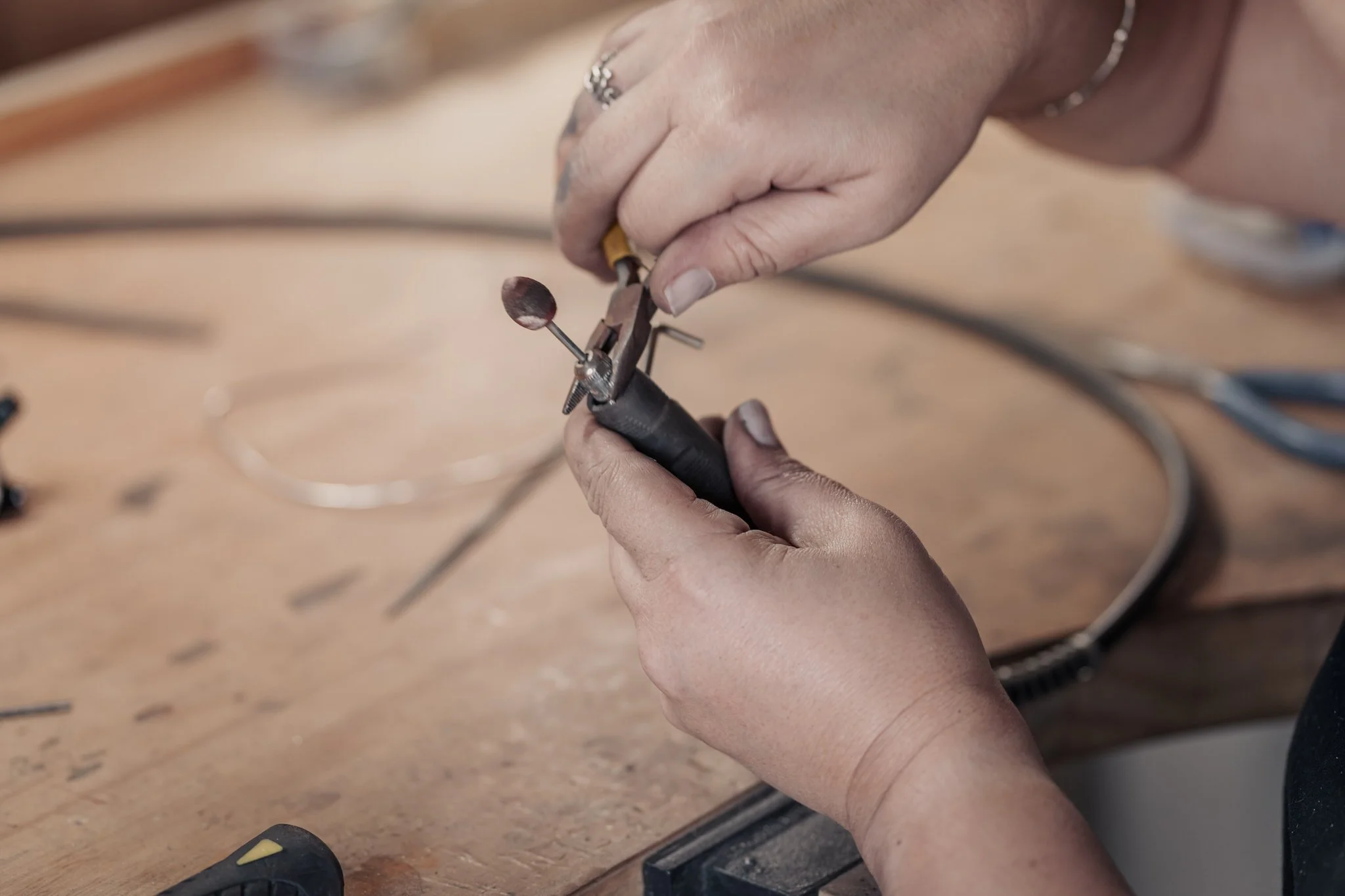 Person holding a small power tool with a sanding attachment, using pliers to adjust a component, on a wooden workbench with various tools in the background.