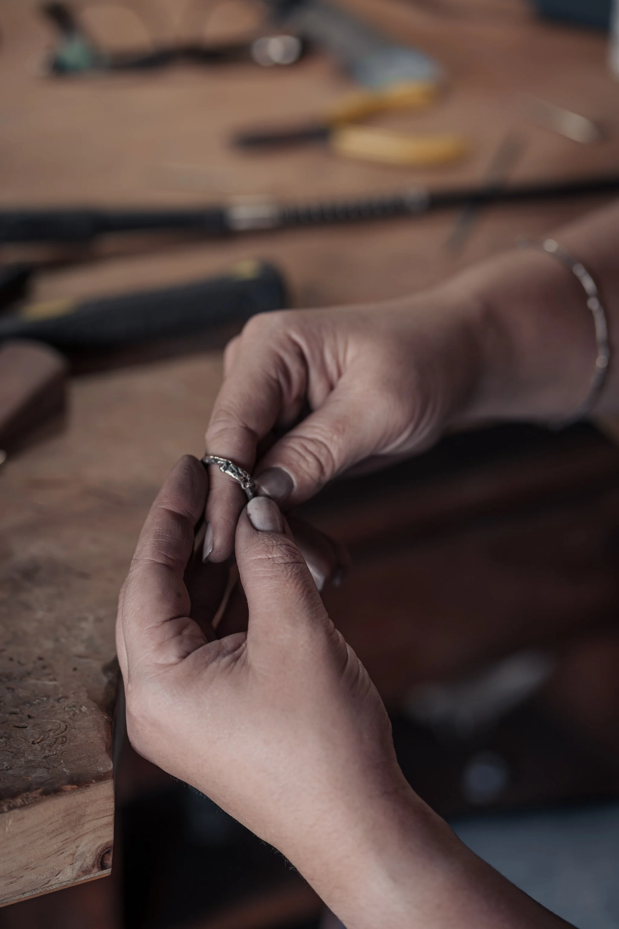 Person holding a small metal object, possibly crafting jewelry, at a workbench with tools in the background.
