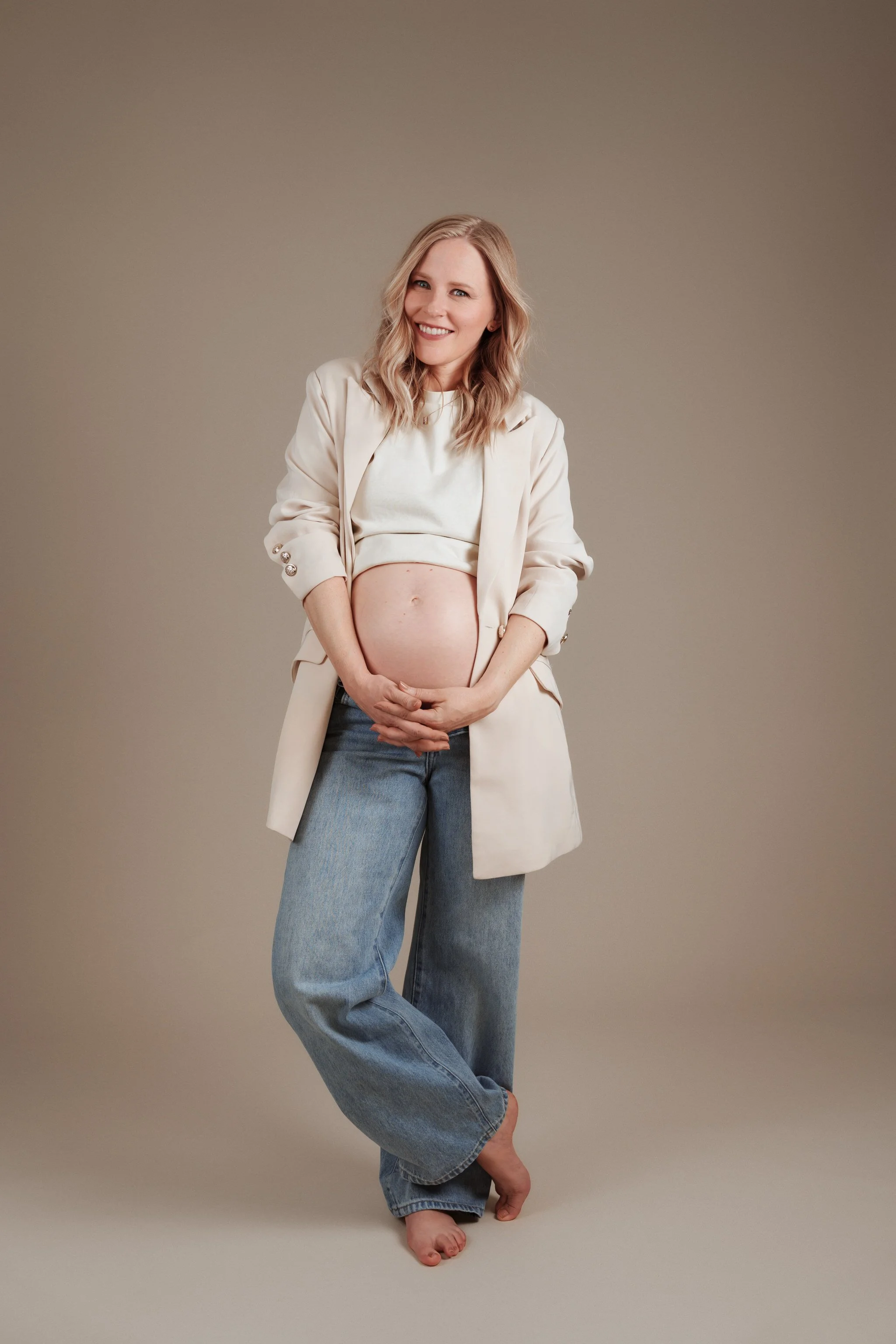 A woman with blonde hair smiling, dressed in a light-colored blazer and jeans, holding her pregnant belly, standing barefoot against a beige background.