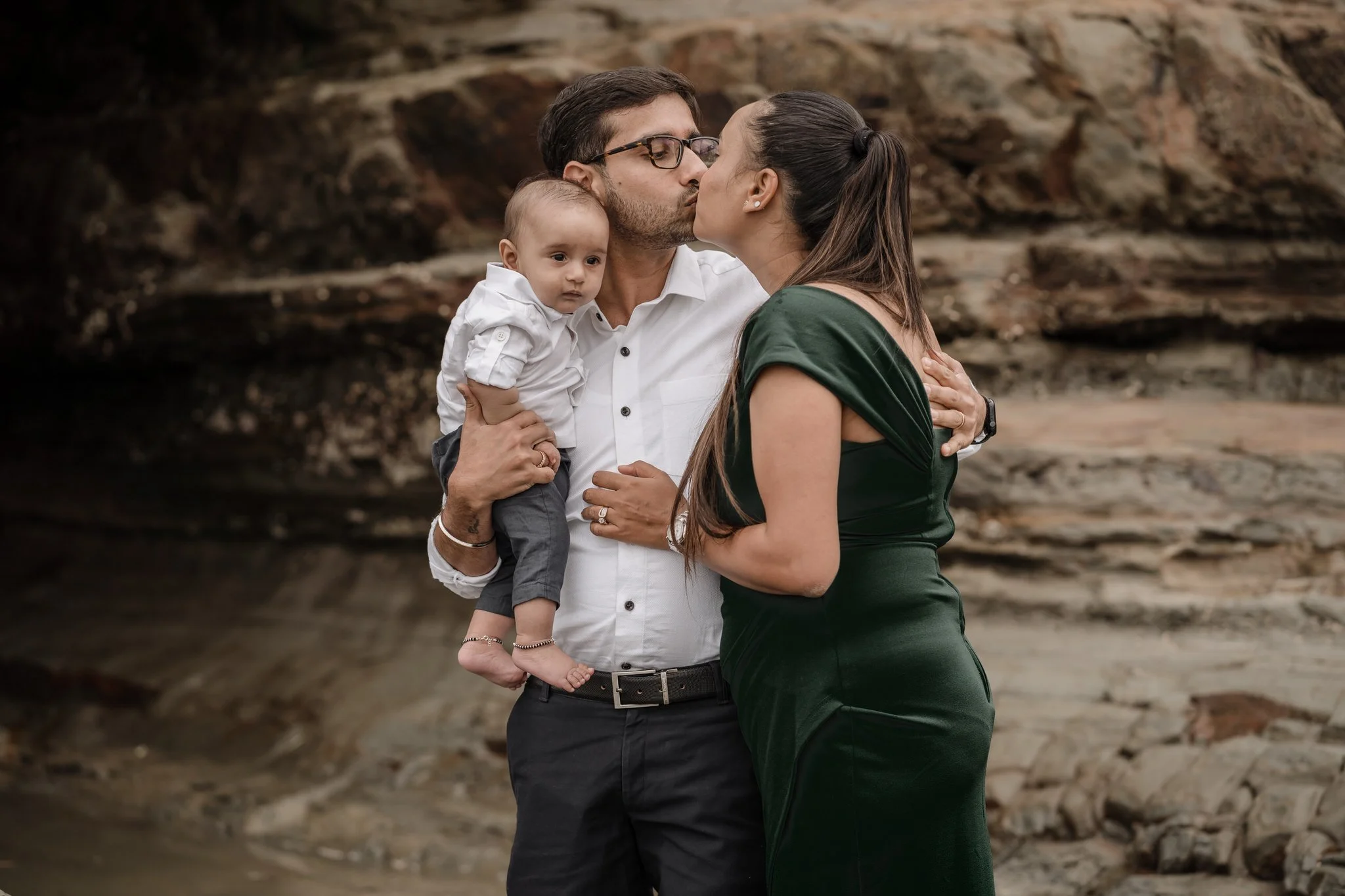 A couple standing outdoors, embracing and kissing, with a man holding a baby in his arms. The woman is wearing a green dress, while the man is in a white shirt. Rocky background.