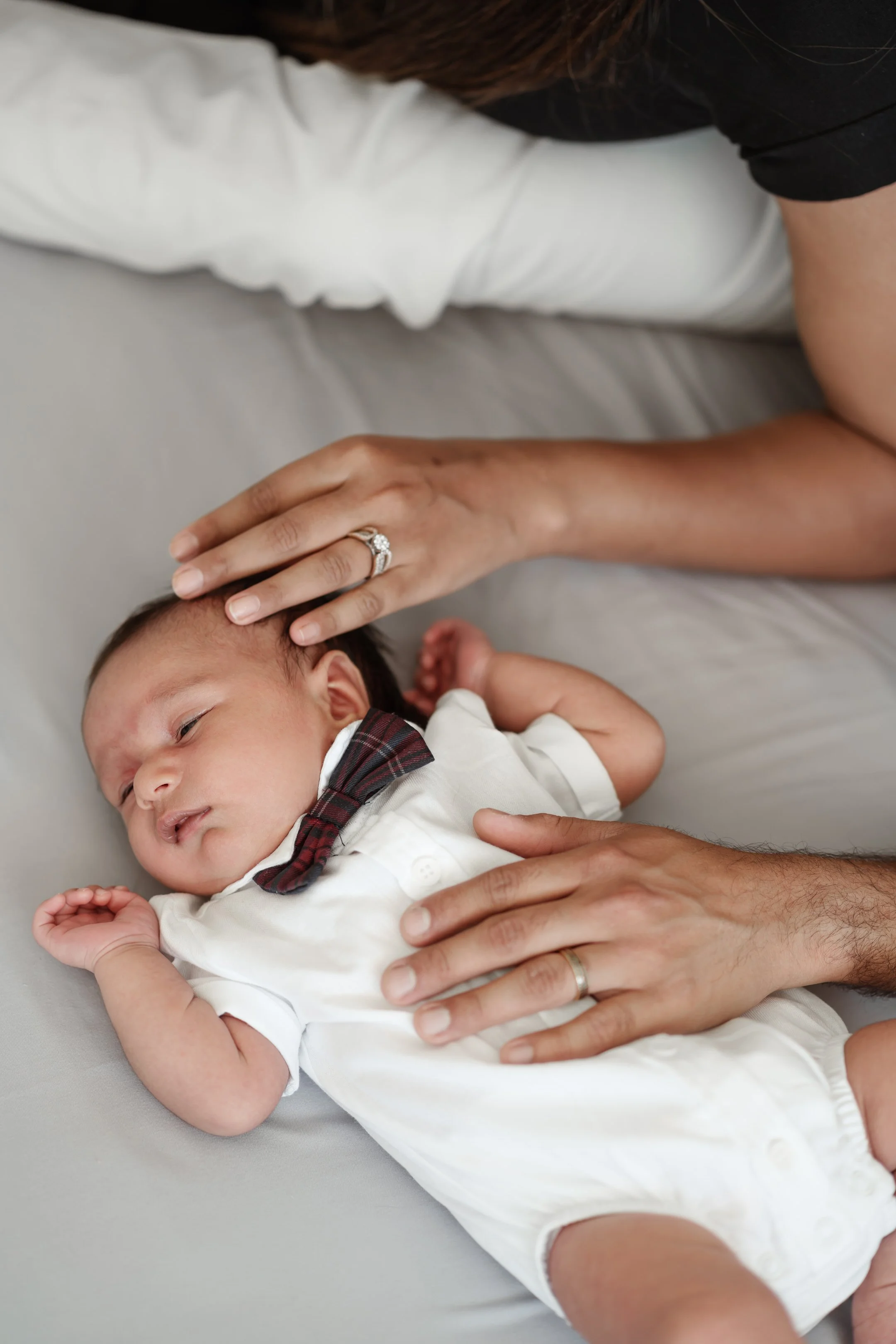 Baby wearing white outfit and red bow tie, lying on bed with hands gently touching.