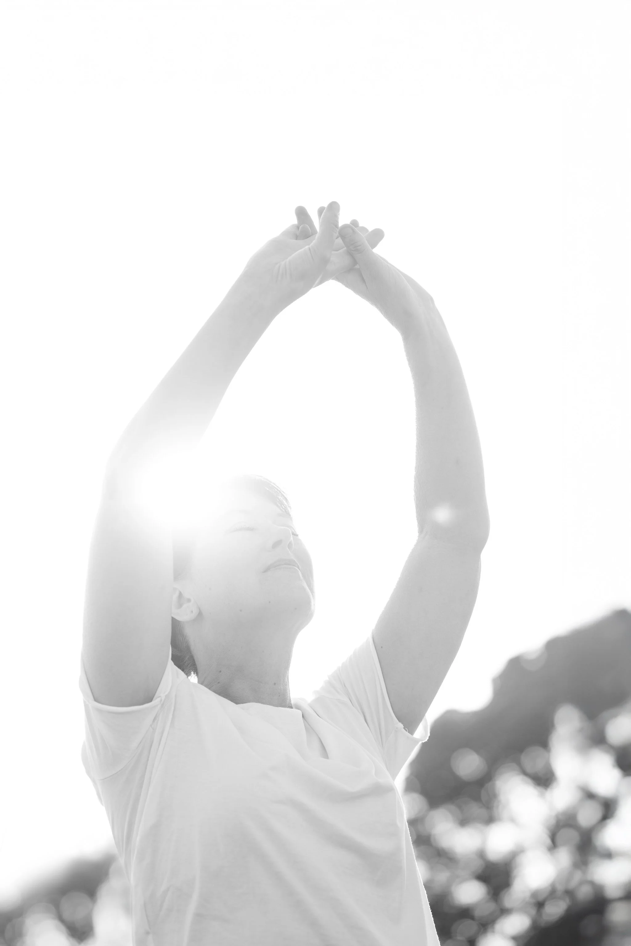A woman in a white shirt is stretching or doing yoga outdoors under bright sunlight, with blurred trees in the background.
