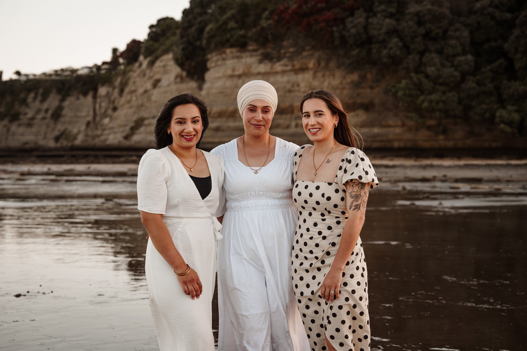 Three women standing together on a beach with cliffs and trees in the background, wearing dresses and smiling.