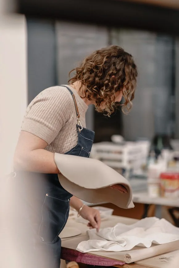 Person with curly hair working on a craft project, holding a piece of fabric, wearing a knit top and overalls, at a work table in a workshop.