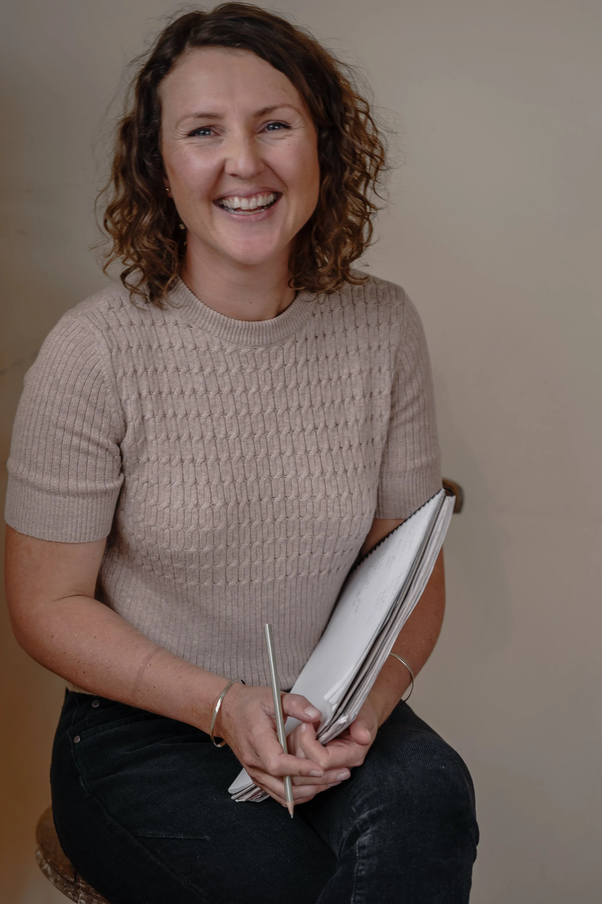 Smiling person holding a notepad and pencil, wearing a beige sweater.