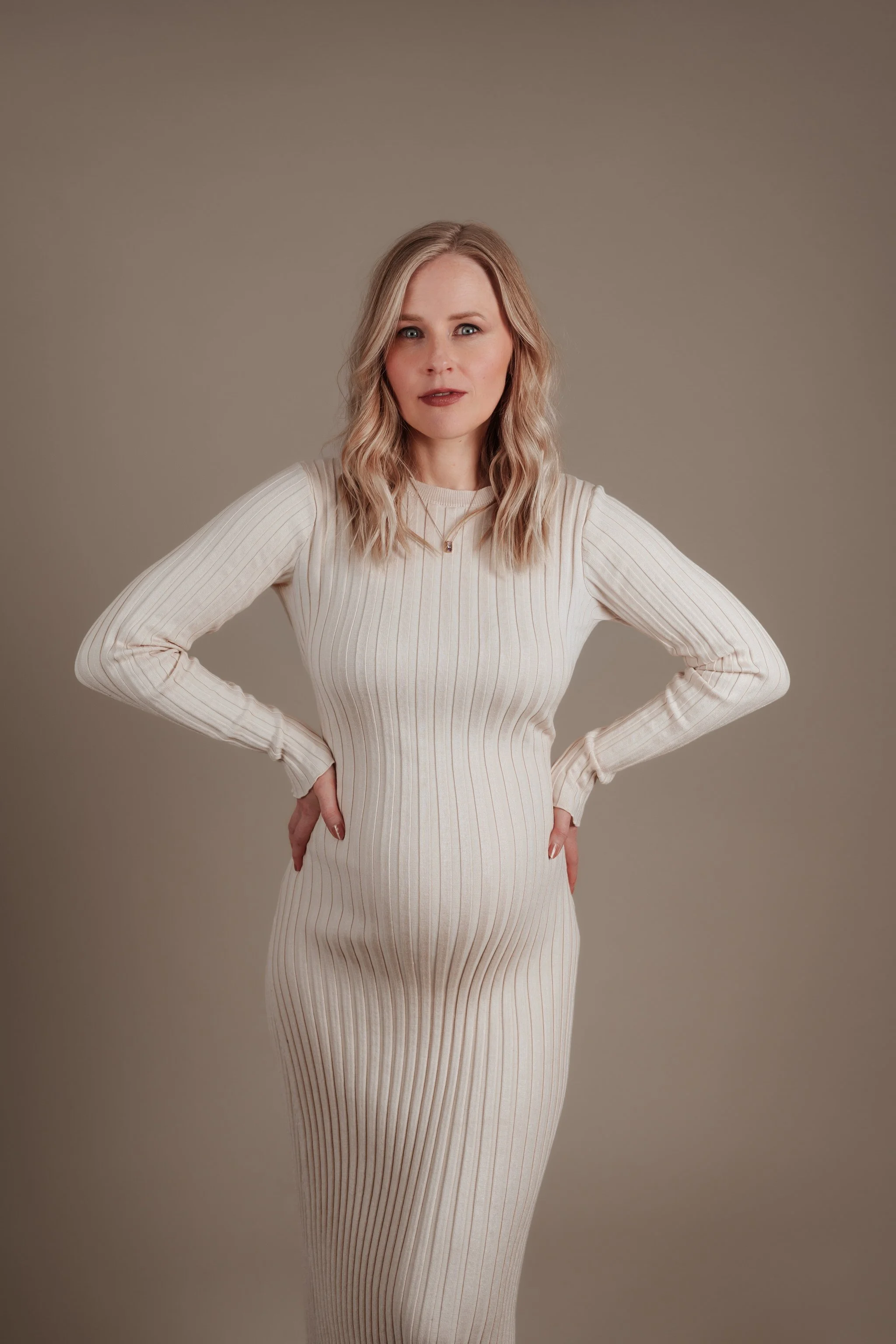 A woman with blonde wavy hair and blue eyes wears a cream-colored, long-sleeve, ribbed dress and poses with her hands on her hips against a neutral background.