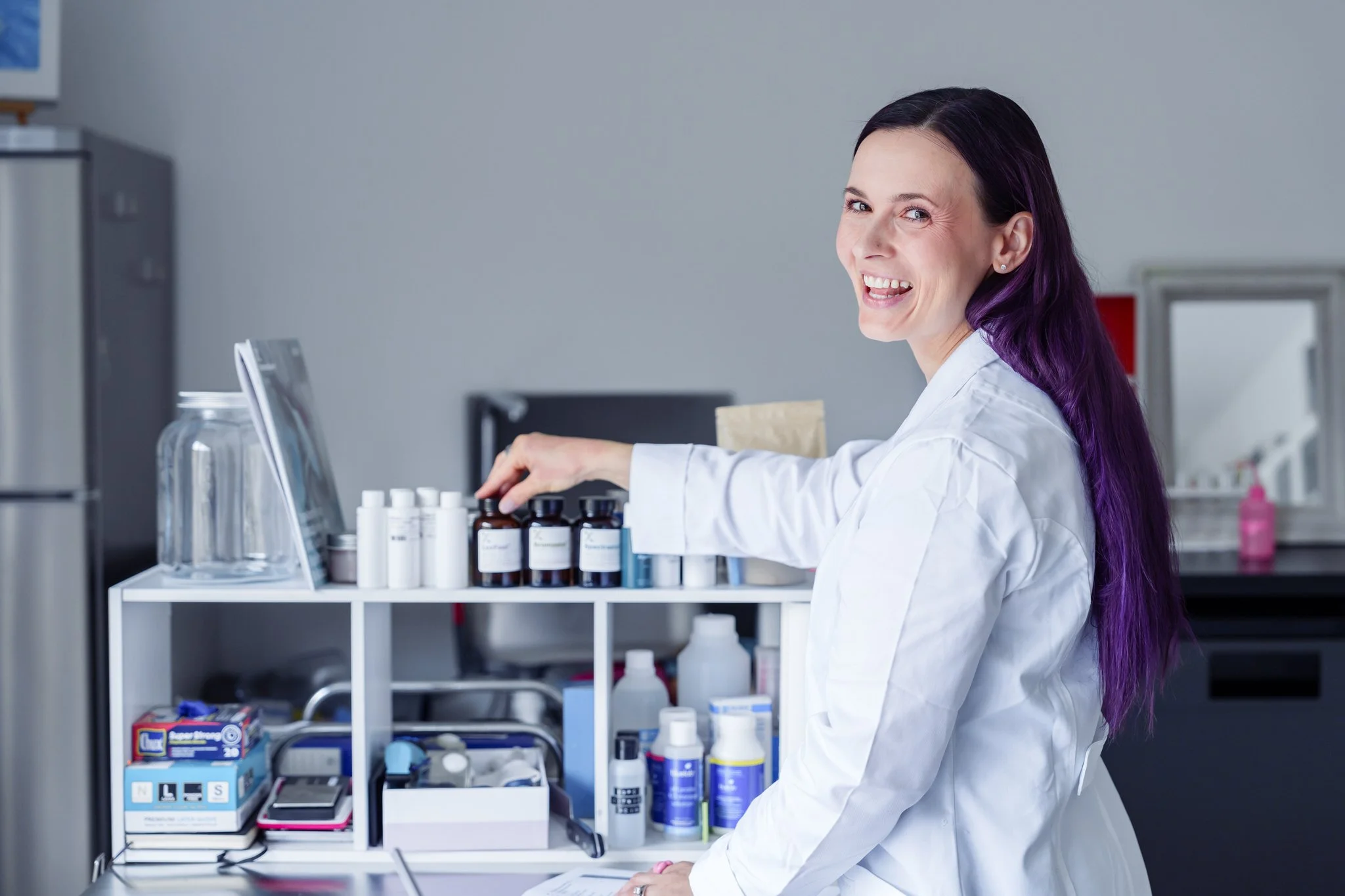 Woman in lab coat smiling, organizing products on a shelf in a laboratory or workspace, with bottles and jars arranged neatly.