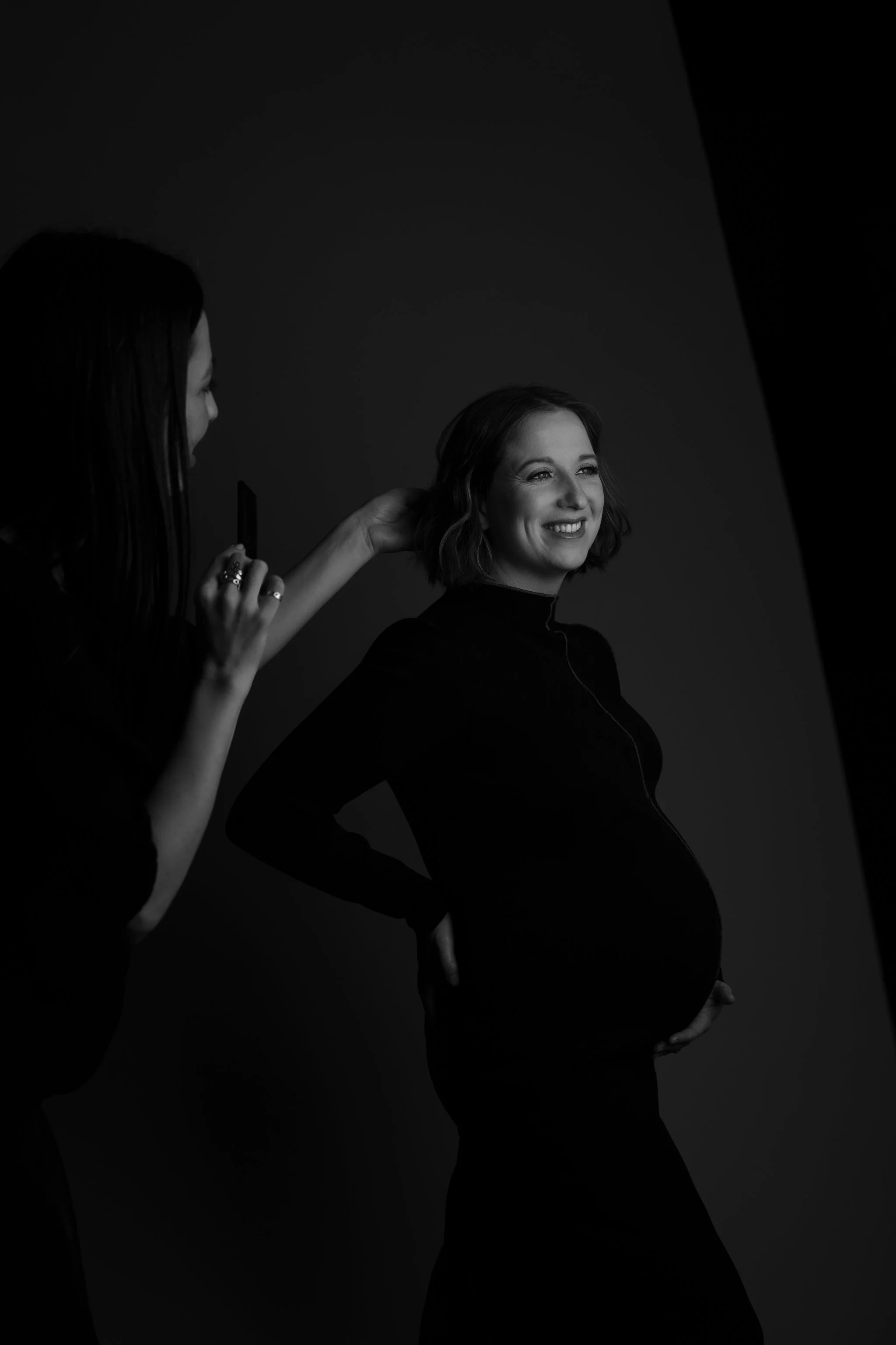 Black and white photo of a pregnant woman smiling, with her hand on her belly, during a photoshoot. A photographer is adjusting her hair.